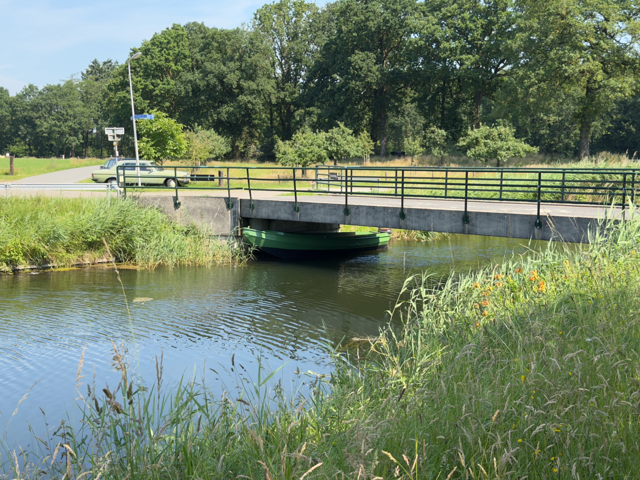 A small footbridge crossing a canal beside a road, with a green rowing boat moored beneath it.
