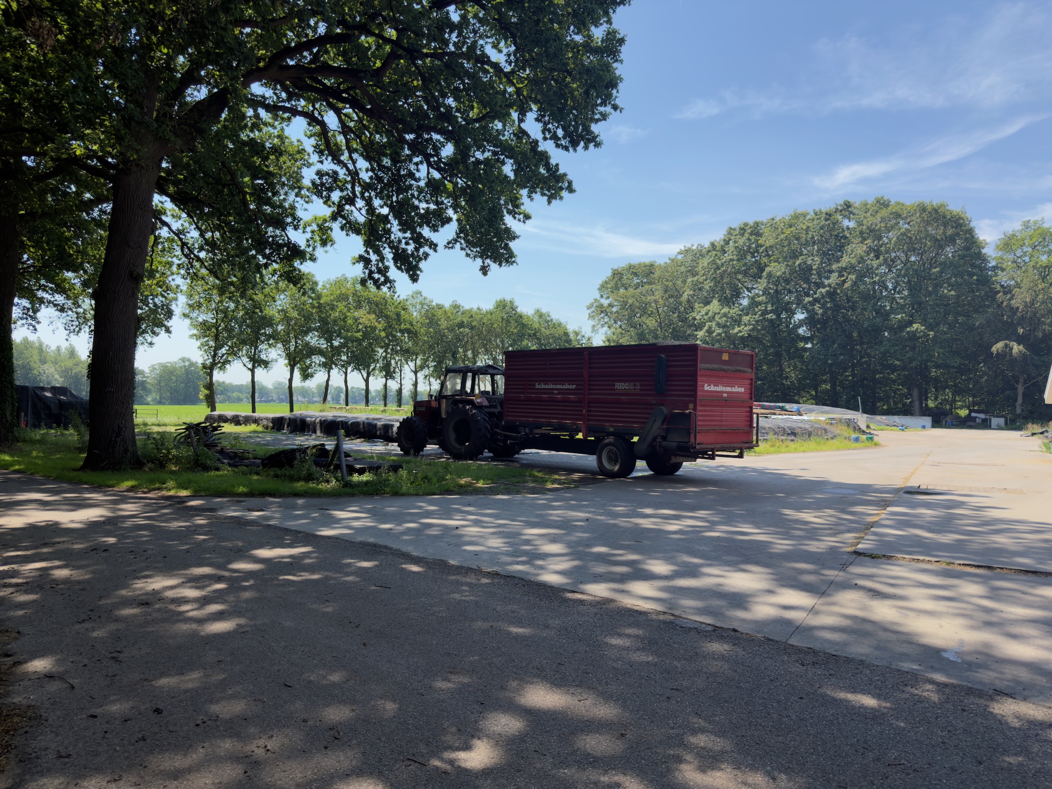 A red tractor and trailer parked under a tree beside a rural road.