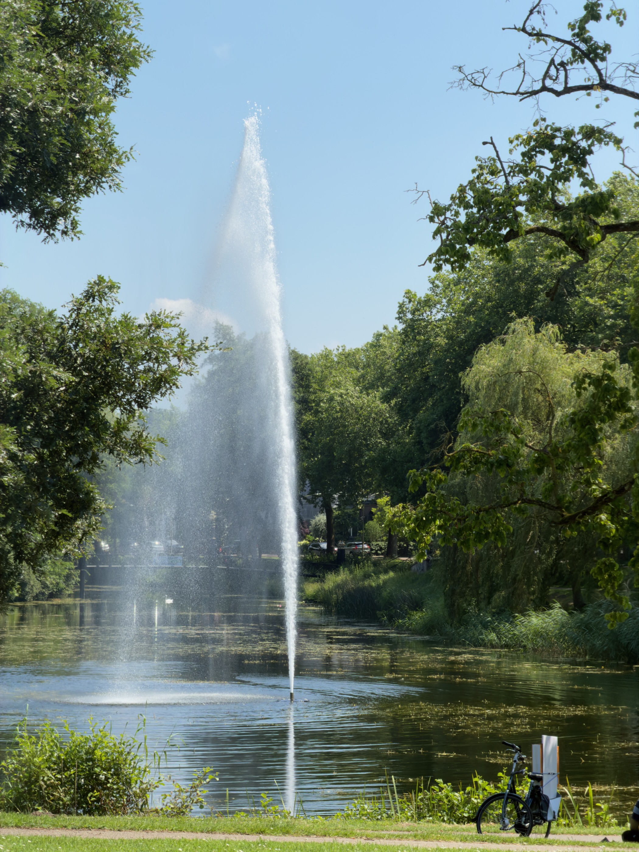 A tall fountain spraying from a park pond, with a bicycle parked on the bank.