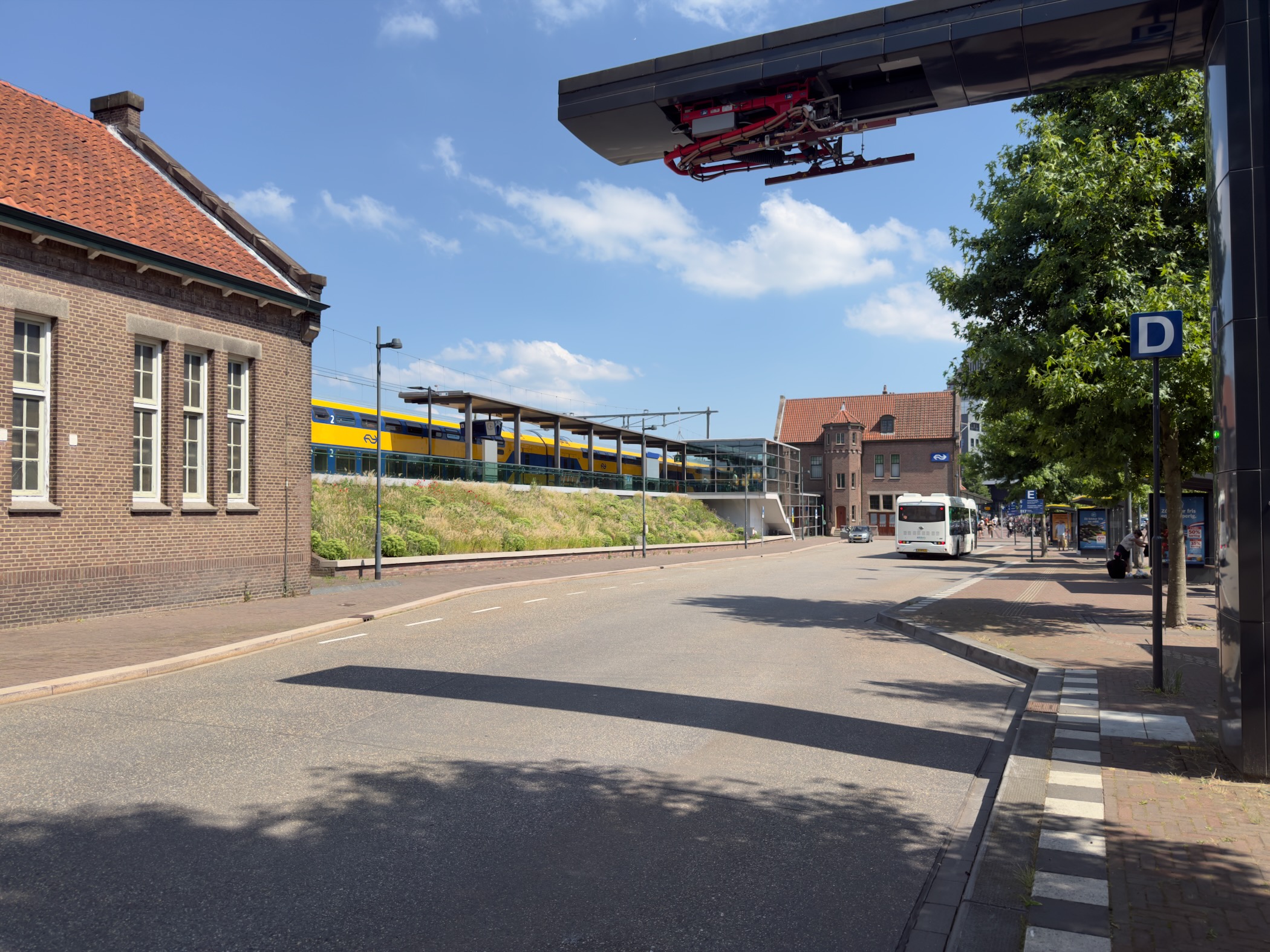 A street by Deventer station with a yellow and blue train, an old brick building and a bus stop.