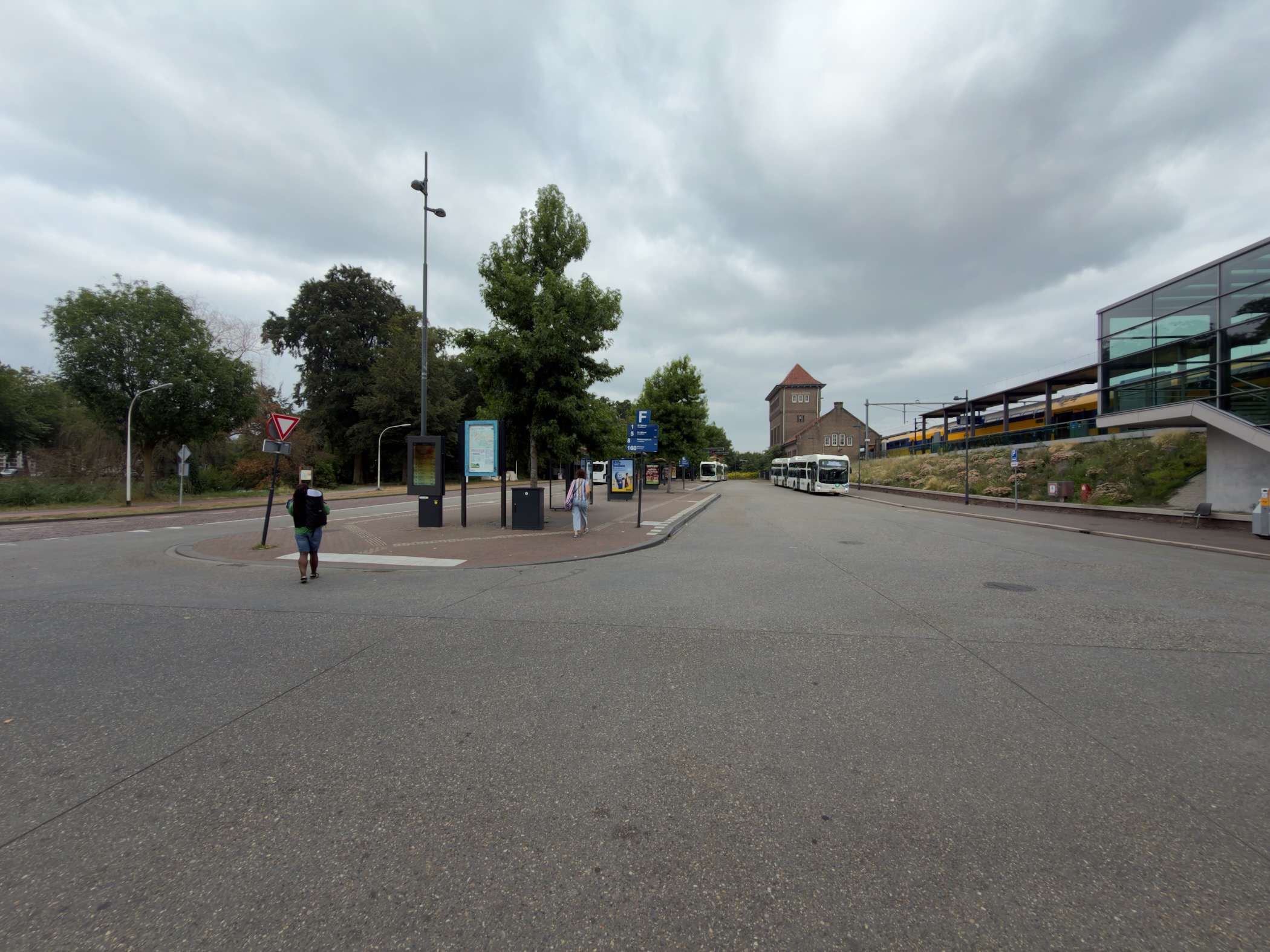 The forecourt of Deventer station with a yellow train, white buses, a brick water tower and overcast sky.