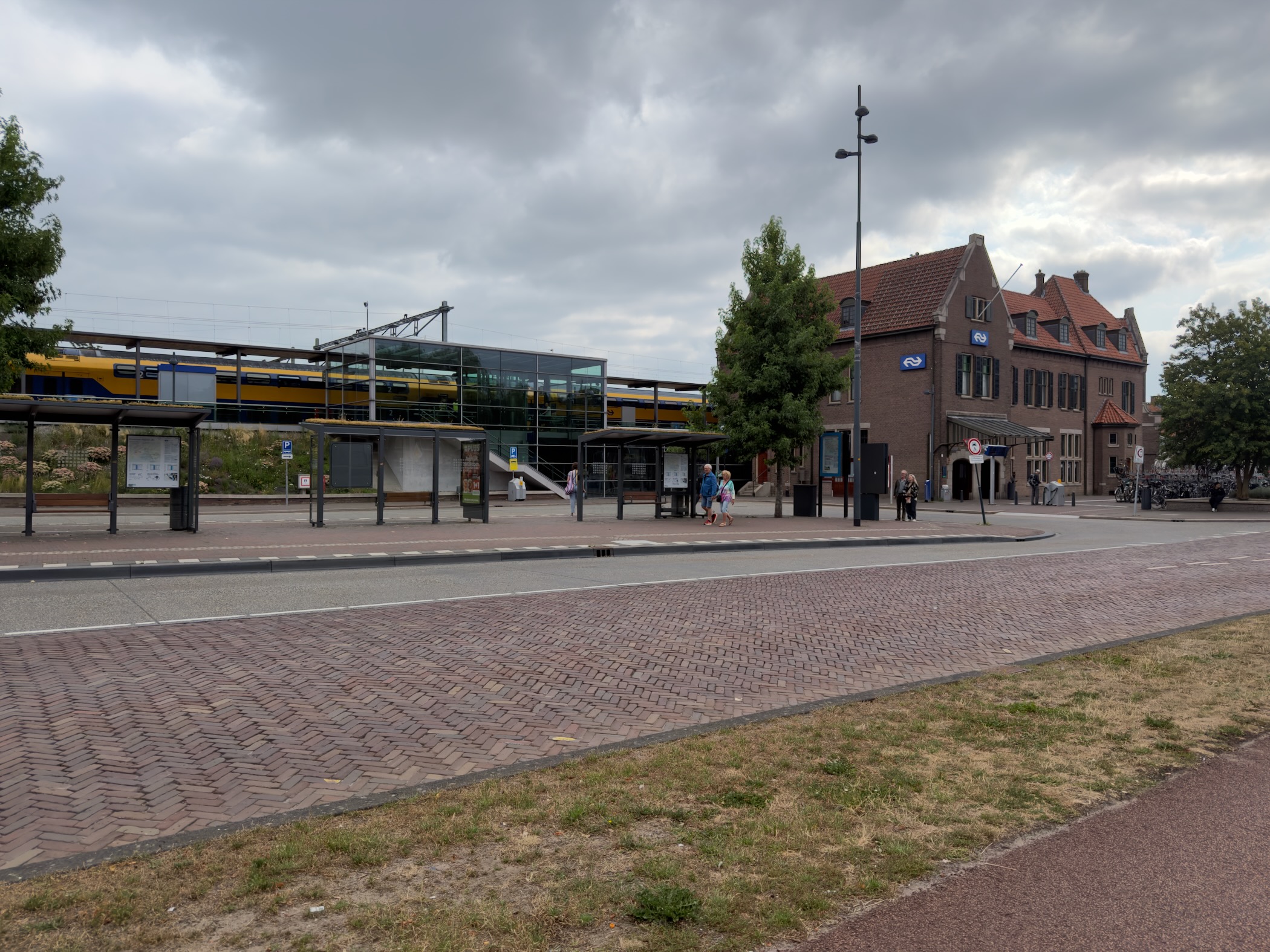The brick Deventer station building with NS logos beside a modern glass canopy and a yellow double-decker train.
