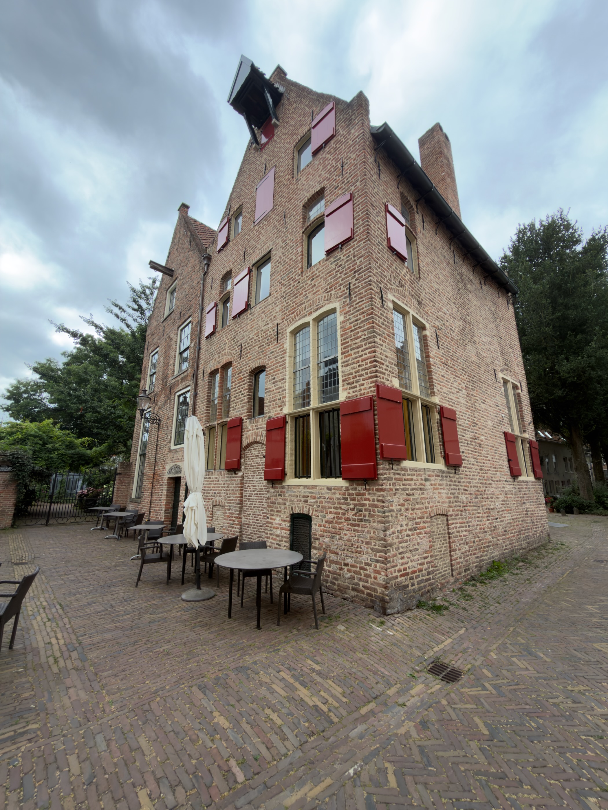 A tall historic brick house with red shutters and leaded windows on a cobbled square with outdoor café tables.