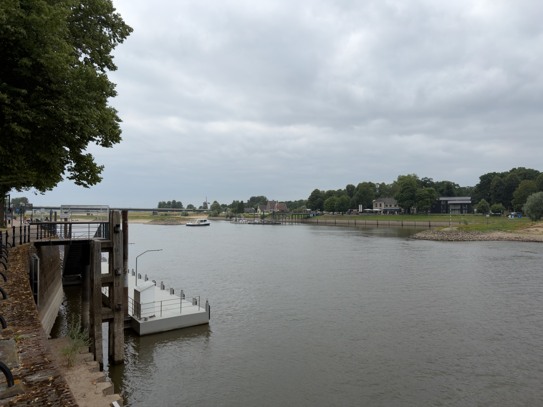 The wide IJssel river from the Deventer quay, with a jetty, distant windmill and bridge under grey cloud.