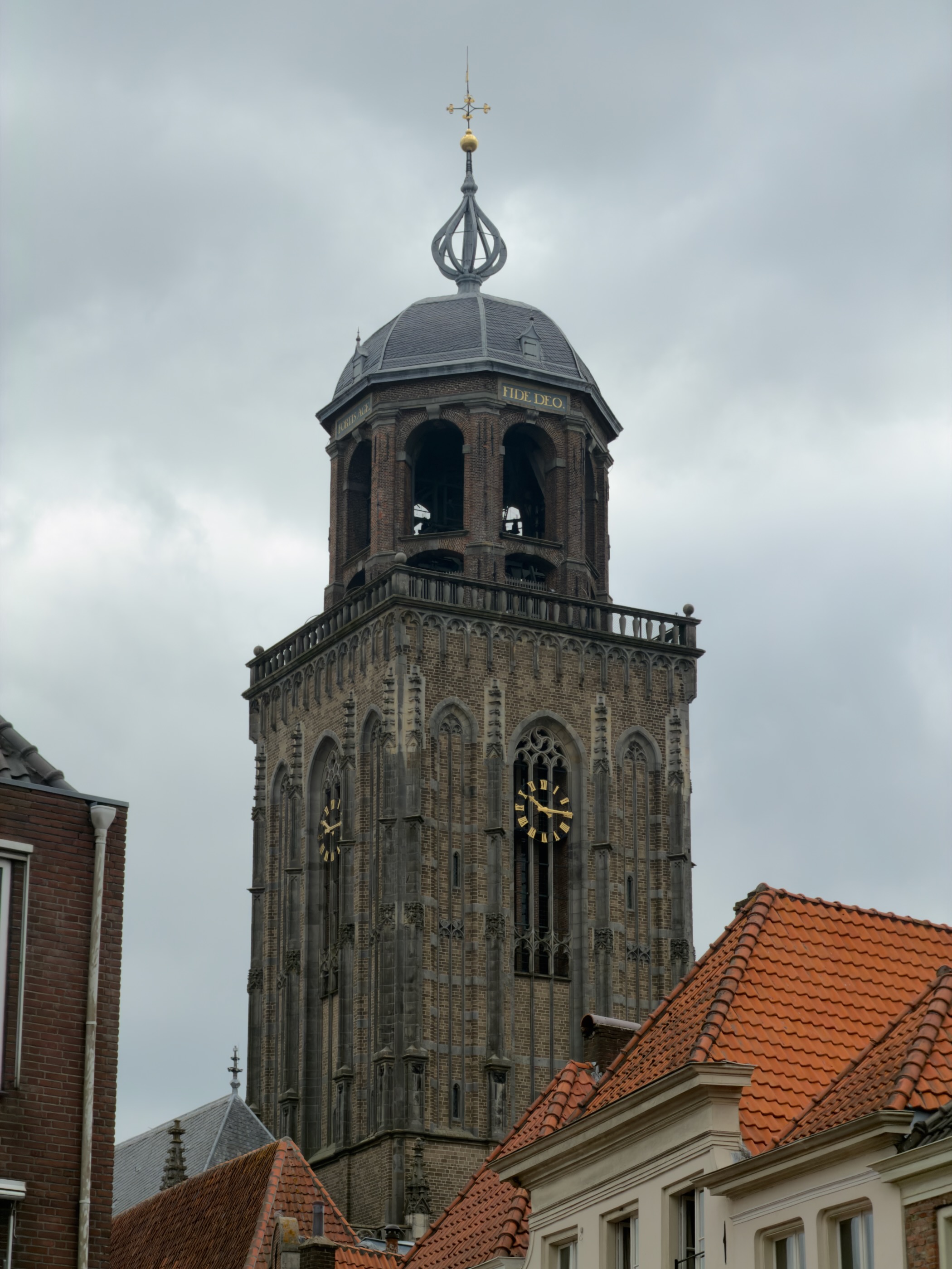The Gothic brick tower of the Lebuinuskerk with gold clocks and a Fide Deo cupola rising above red rooftops.