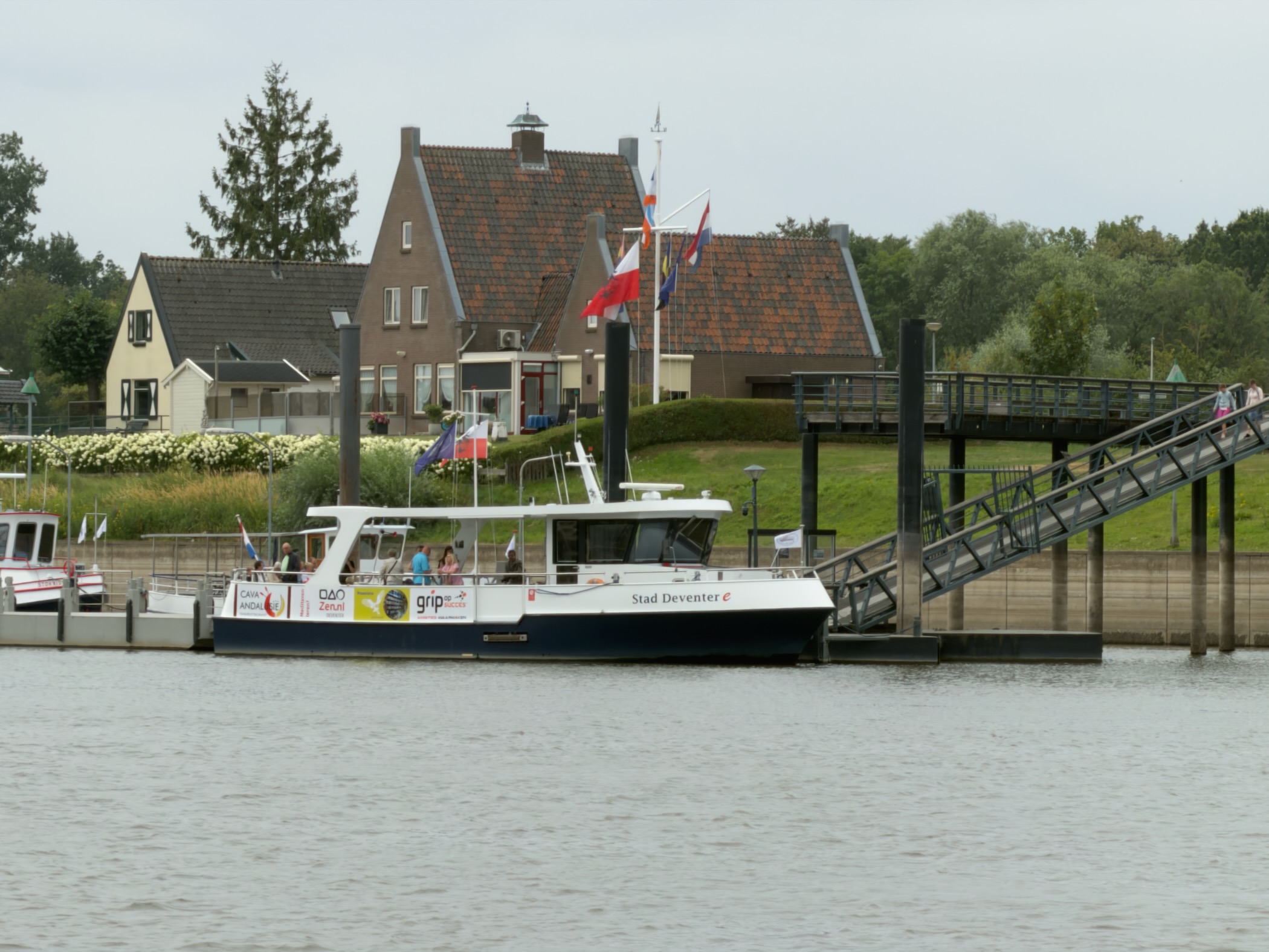 The Stad Deventer ferry moored at a pontoon with a gangway, flags and brick houses with red roofs behind.