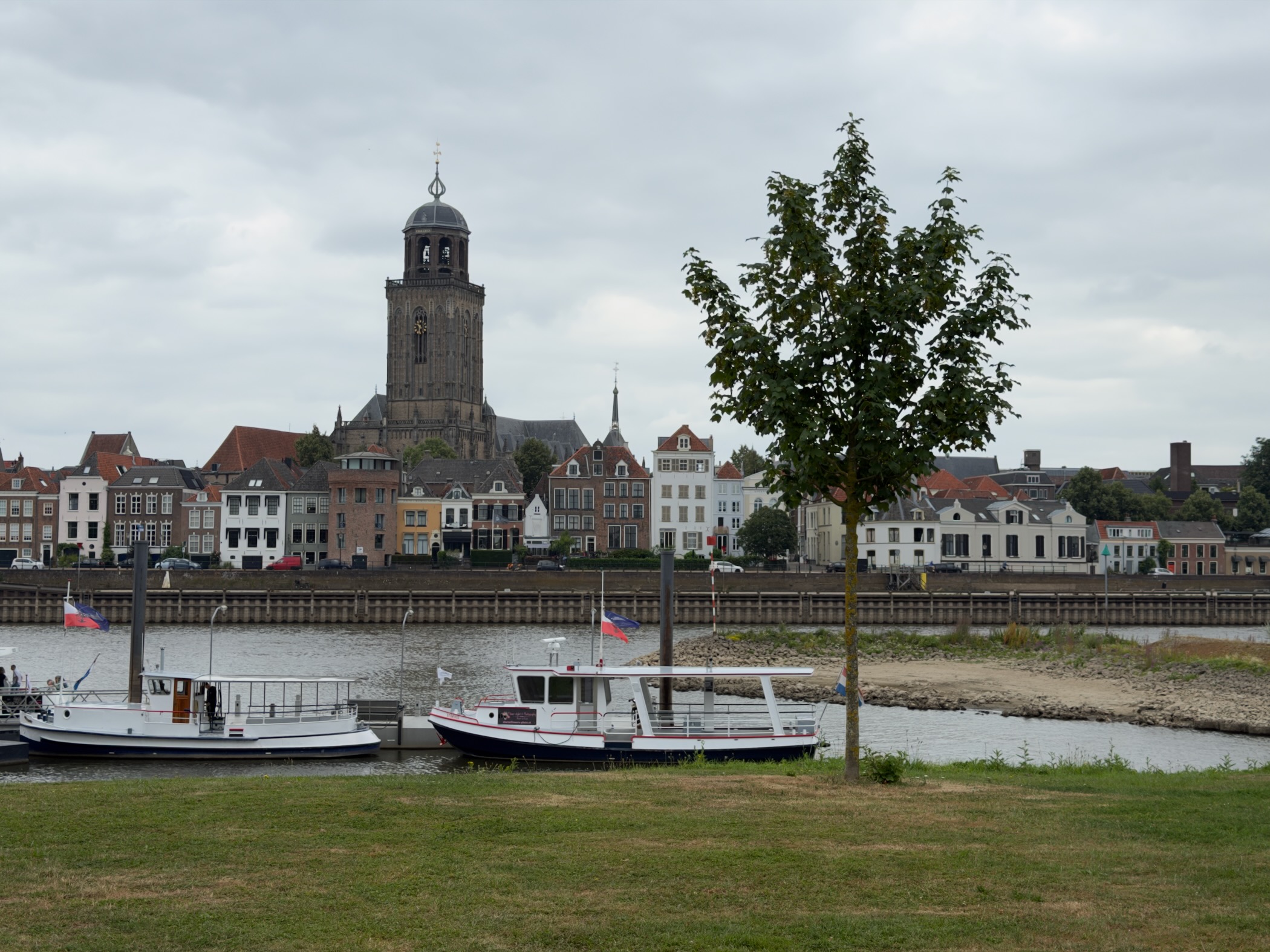 Deventer skyline with the Lebuinuskerk and waterfront houses seen across the IJssel from a grassy bank with two boats.