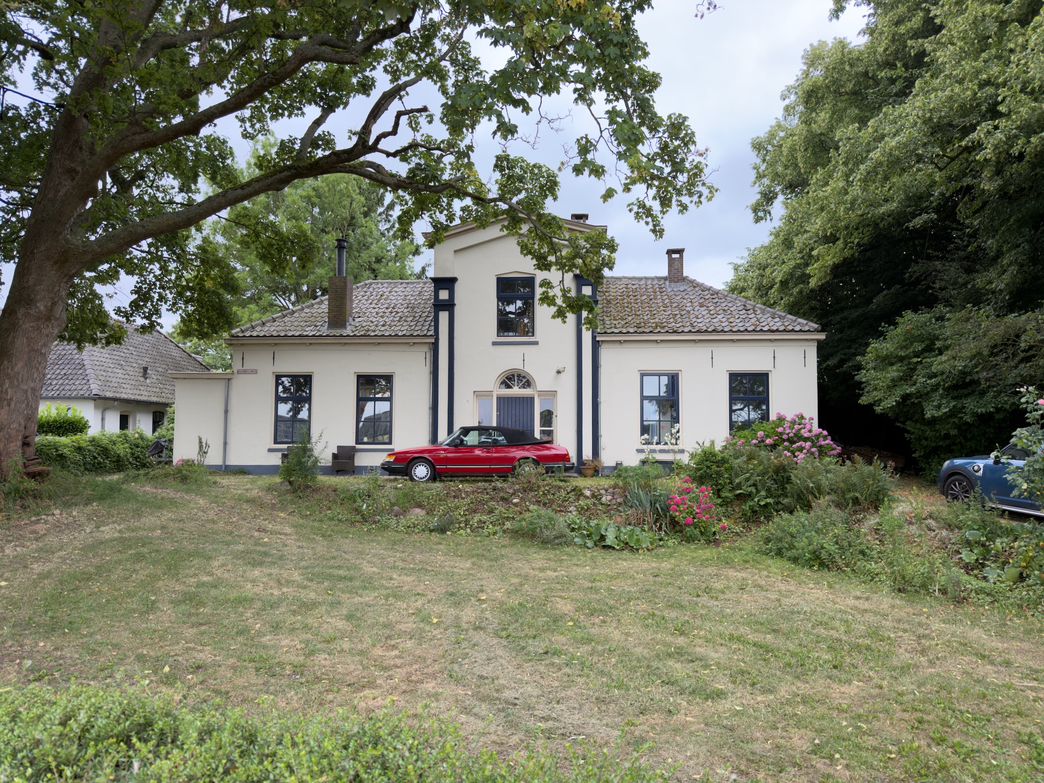 A cream country house with dark blue shutters and a red convertible parked on the lawn under mature trees.