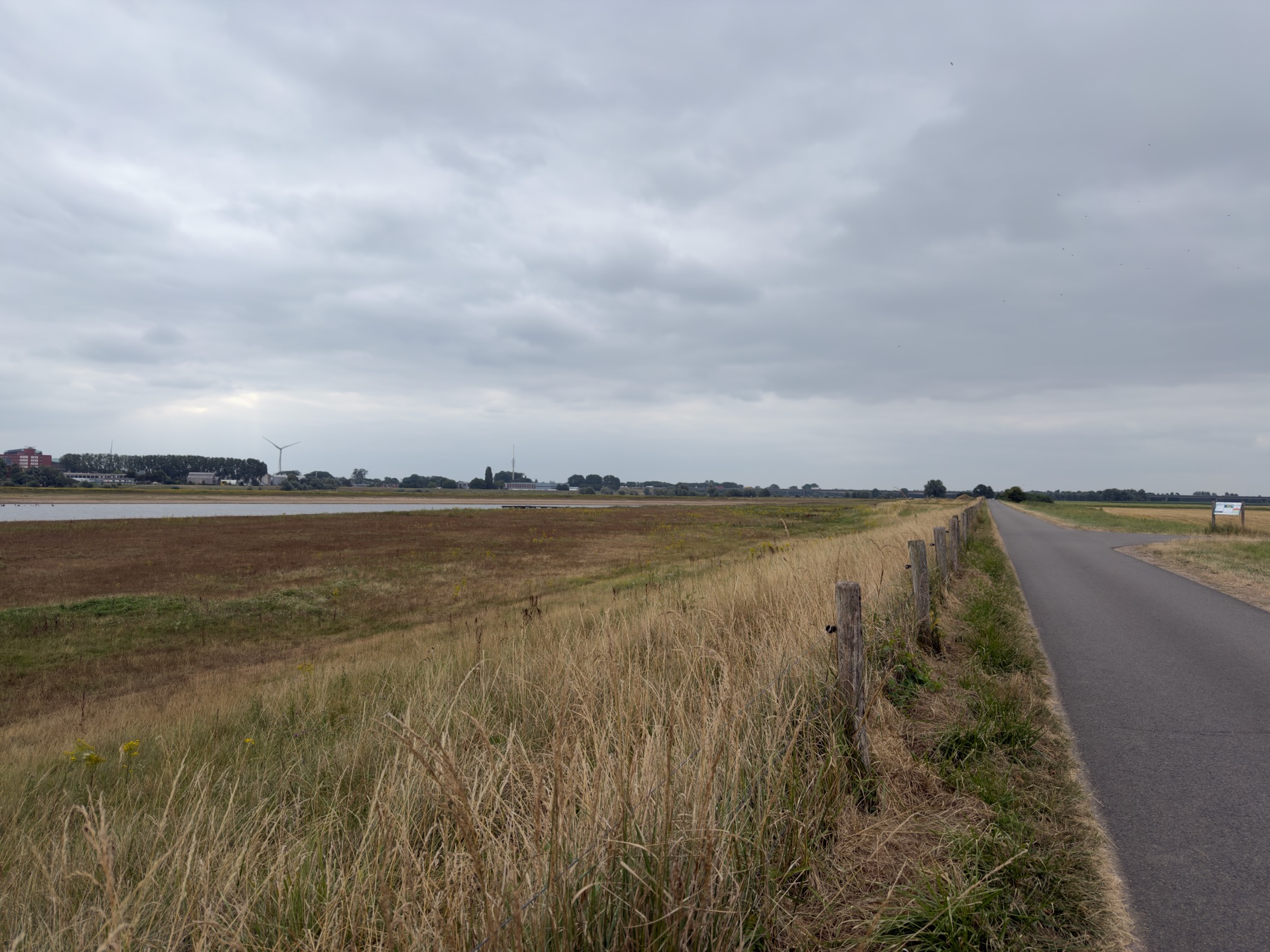 A paved dike path running into the distance between dry floodplain grass and open water under grey cloud.
