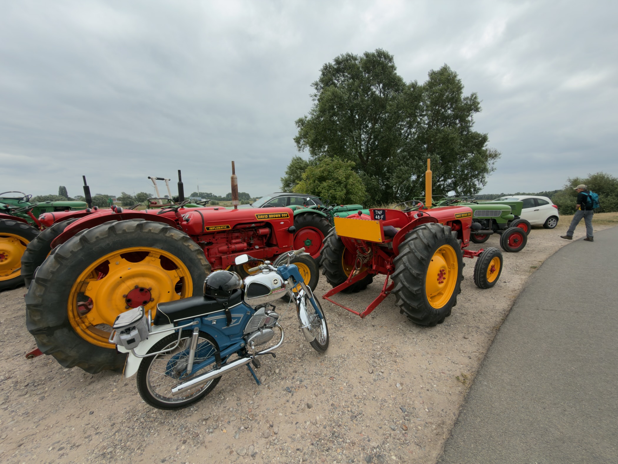 Vintage red and green tractors with yellow wheels parked beside a blue moped on a gravel verge.