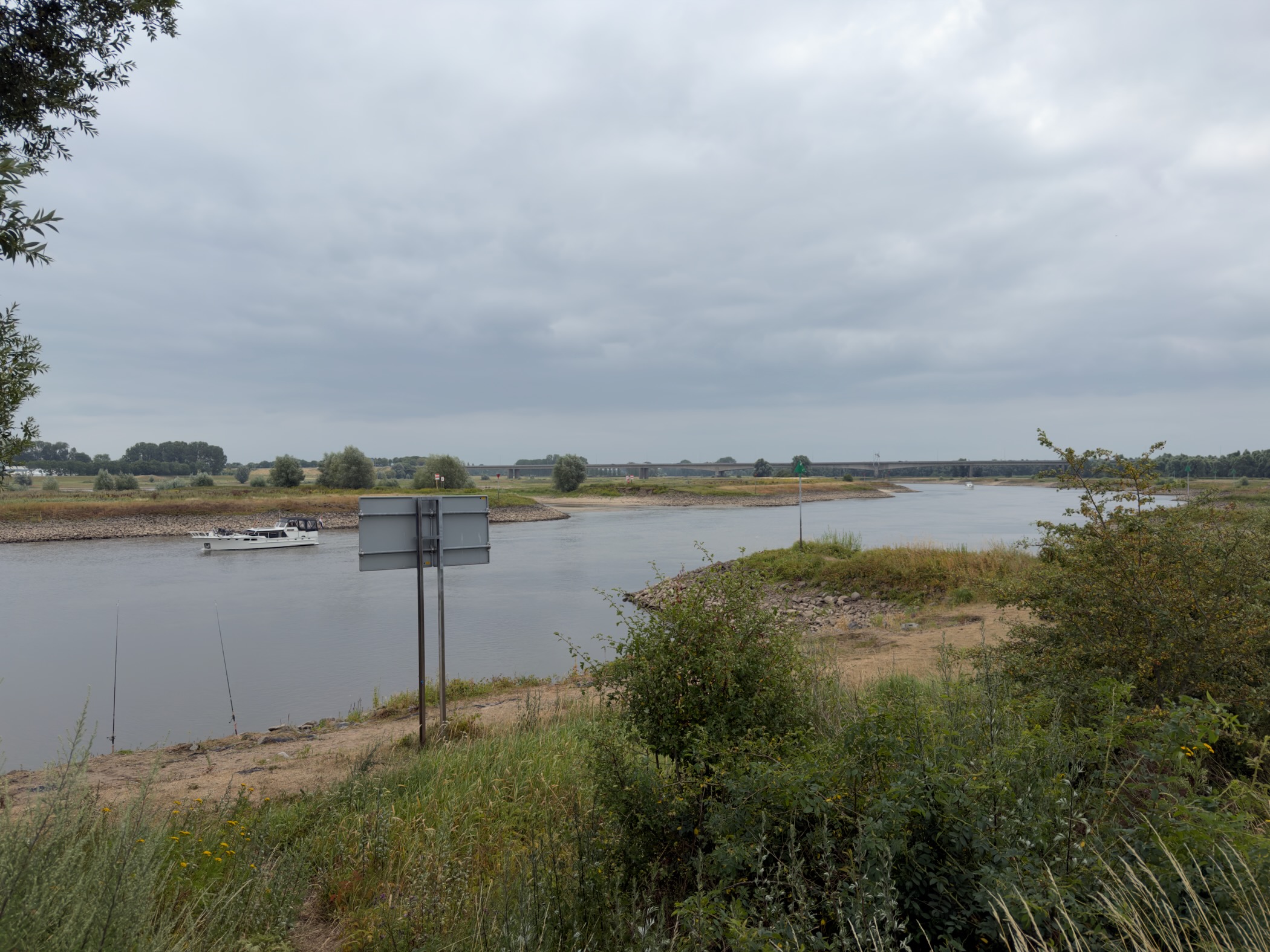 The IJssel curving between rocky banks and bushes, a white boat midstream and a road bridge in the distance.