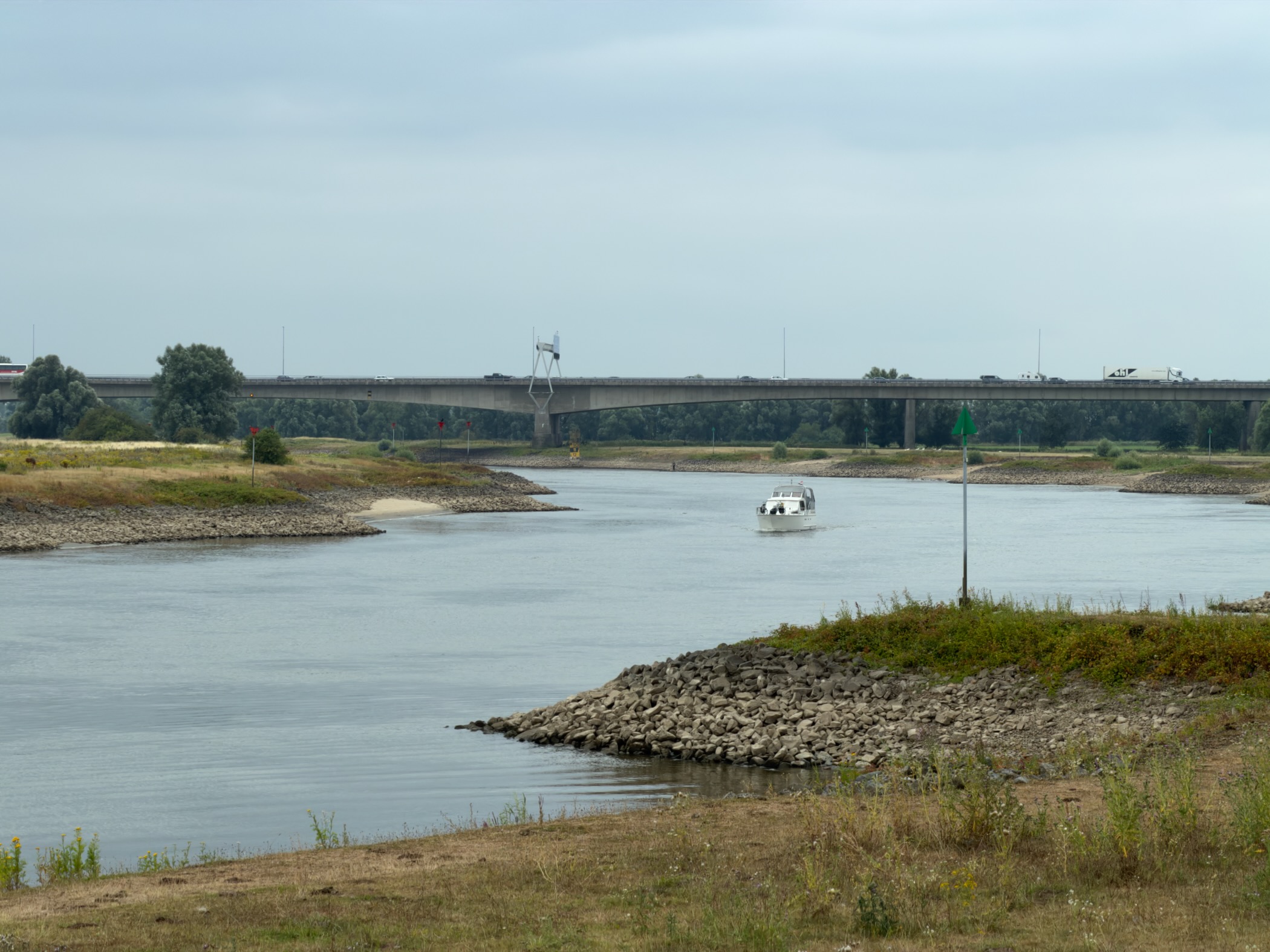 A white motorboat on the IJssel heading towards a long concrete road bridge across the floodplain.