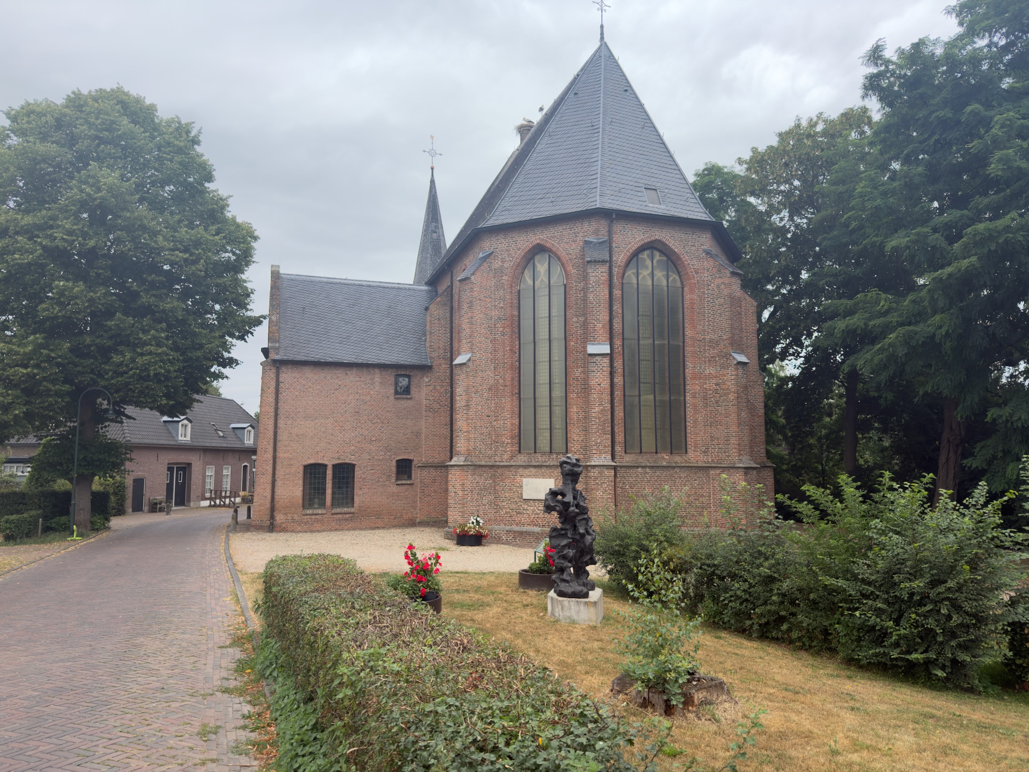The brick apse of Wilp church with tall arched windows, a dark sculpture and flower beds in the grounds.