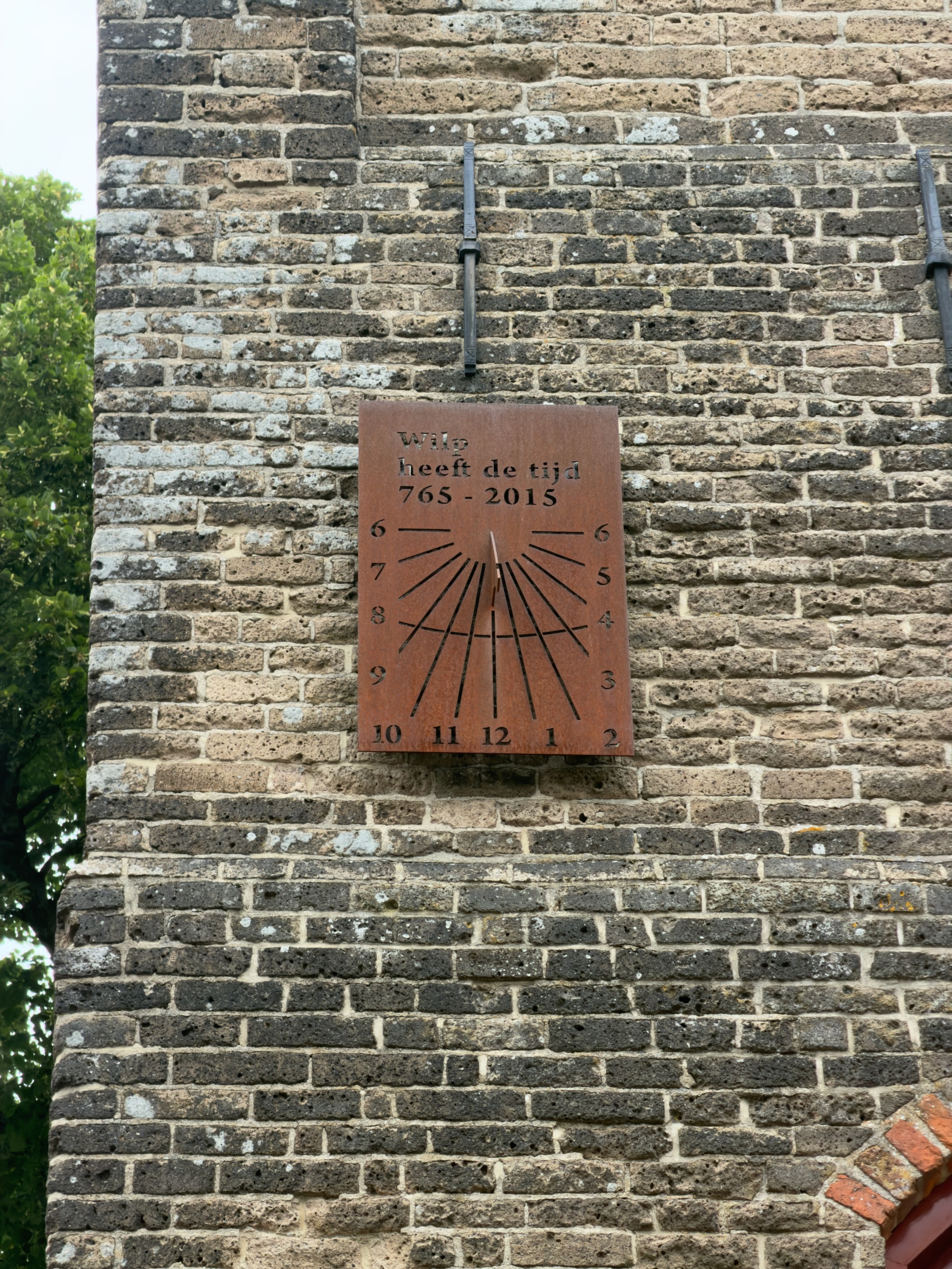 A rusted metal sundial on a brick wall reading Wilp heeft de tijd 765–2015.