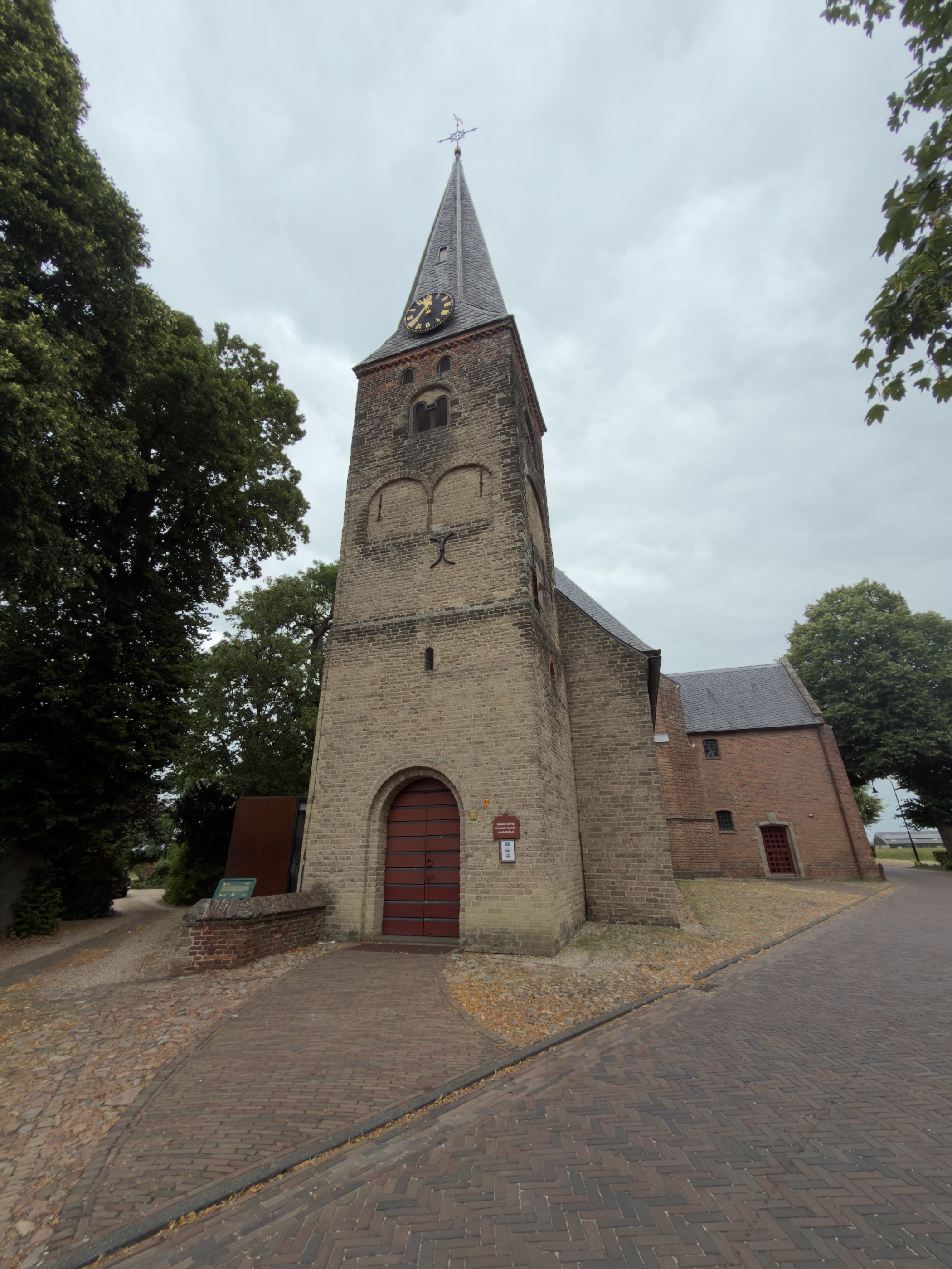 The square brick tower of Wilp church with a pointed spire, clock faces and a red door on a brick path.