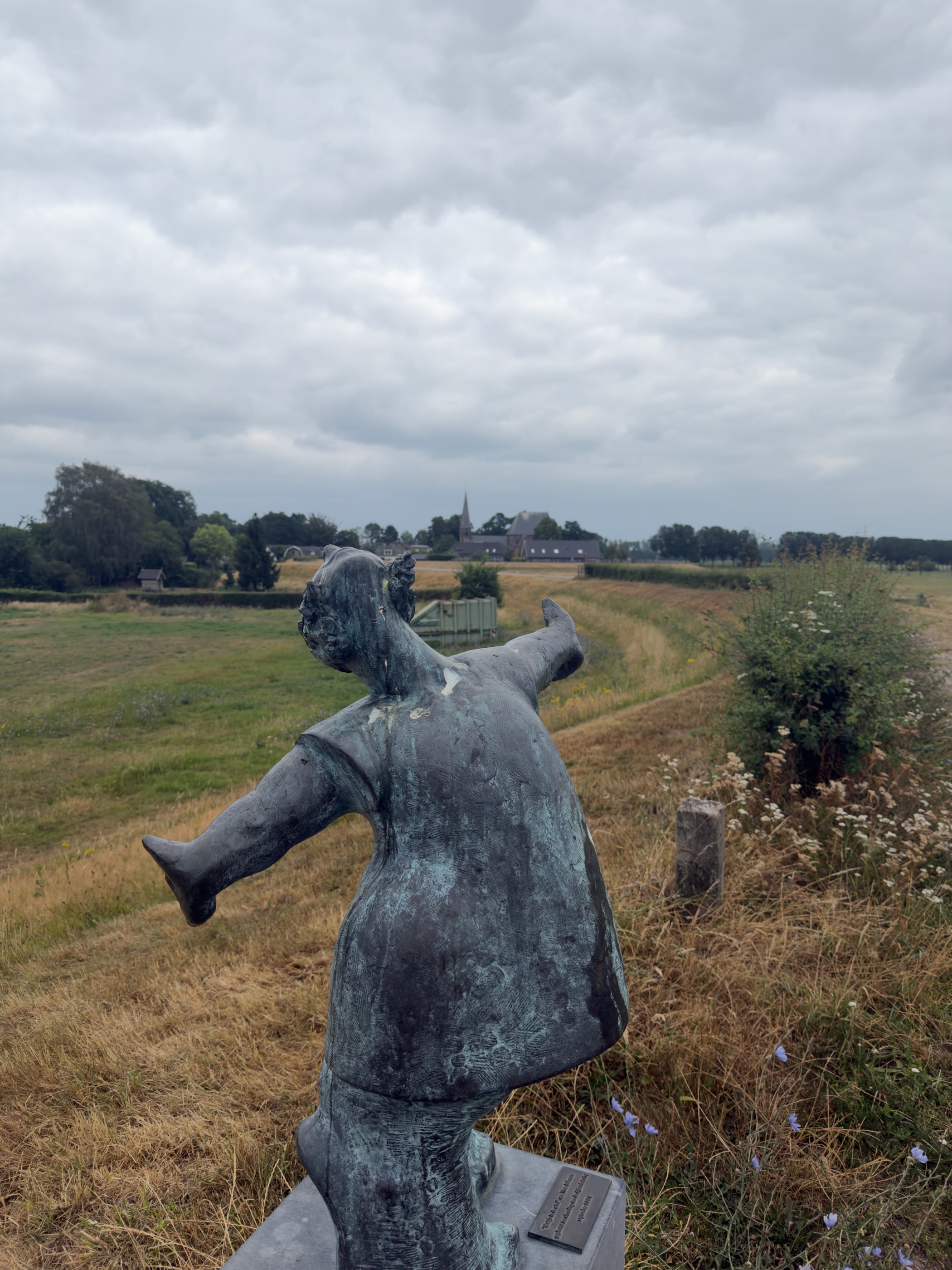 A bronze statue of a plump figure with outstretched arms, looking across fields towards a distant church spire.