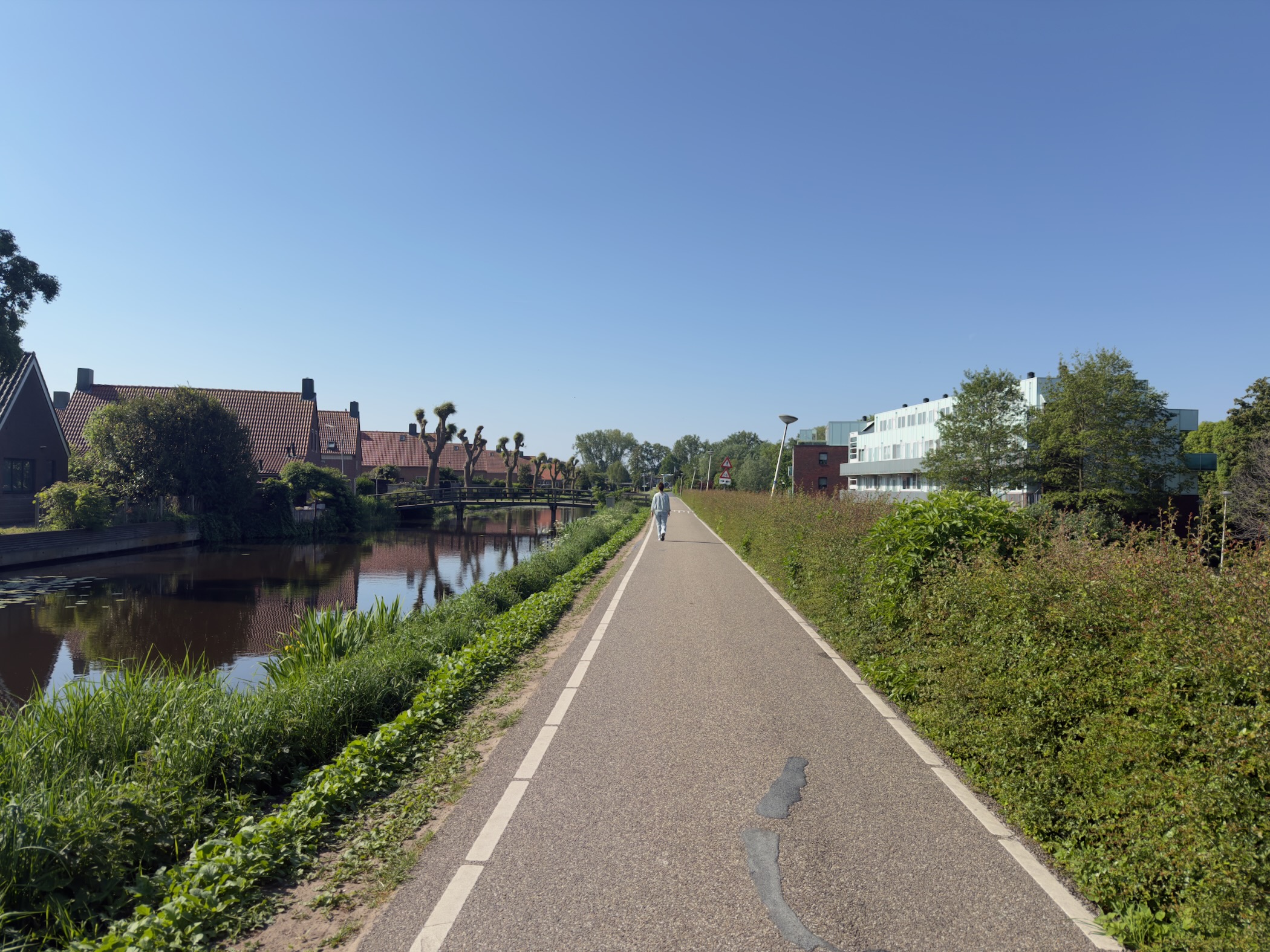 Asphalt path beside a quiet canal with houses on the left and a person walking ahead under a clear blue sky