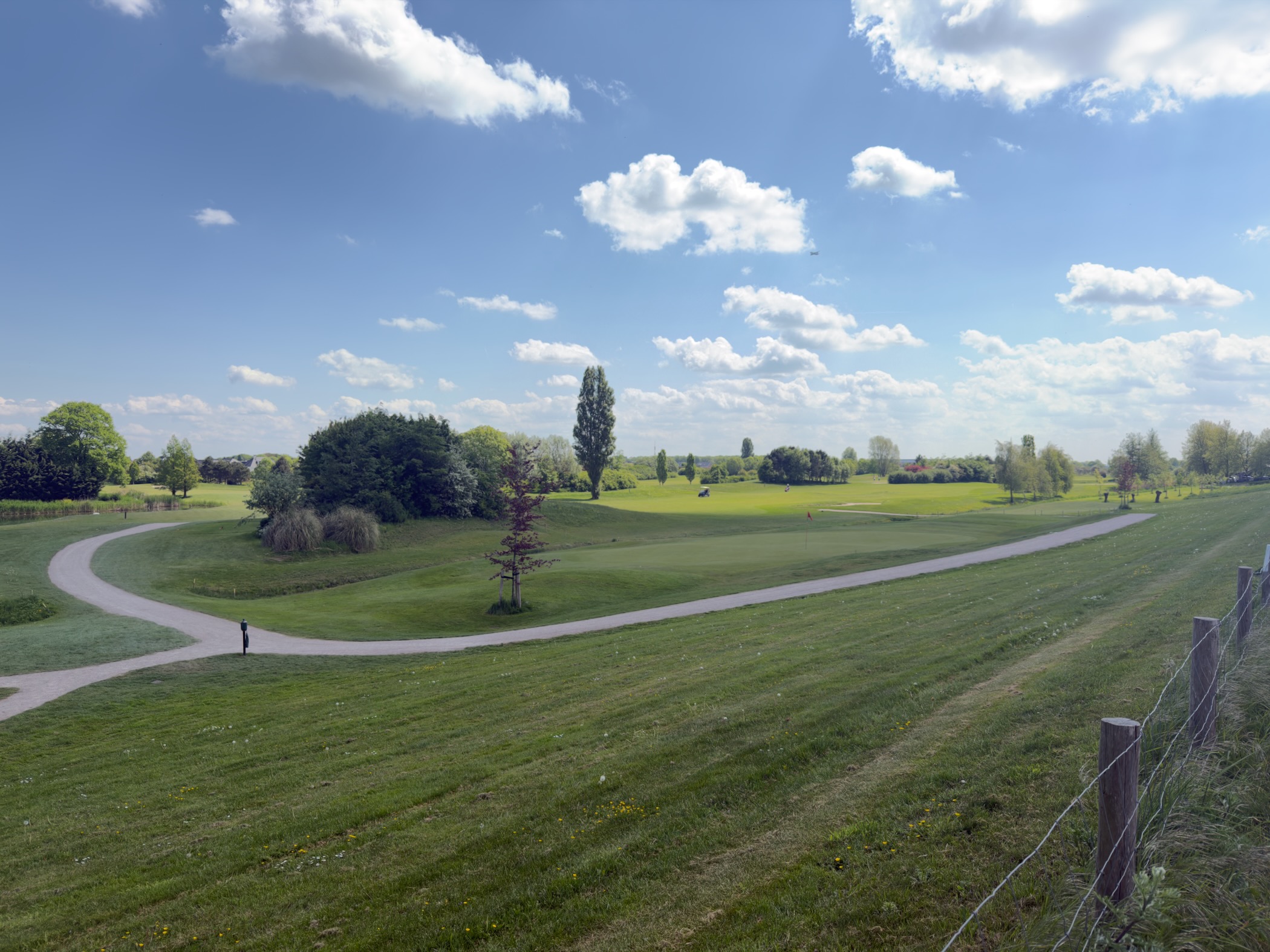 Sweeping view across a golf course with a putting green and red flag under a wide cloud-dotted sky