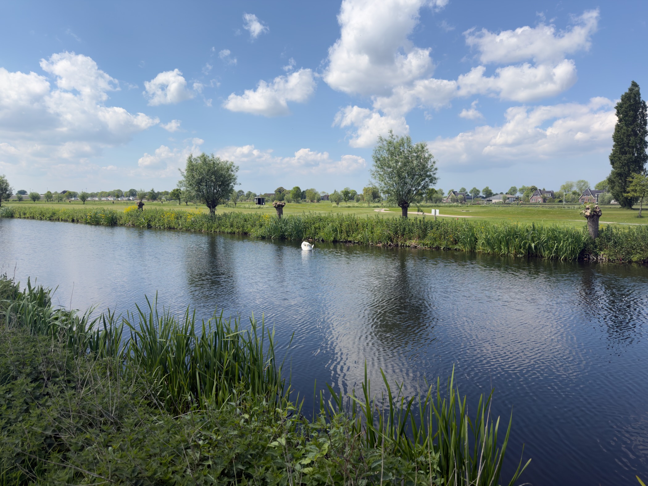 Calm canal flanked by green banks under a bright blue sky scattered with white clouds