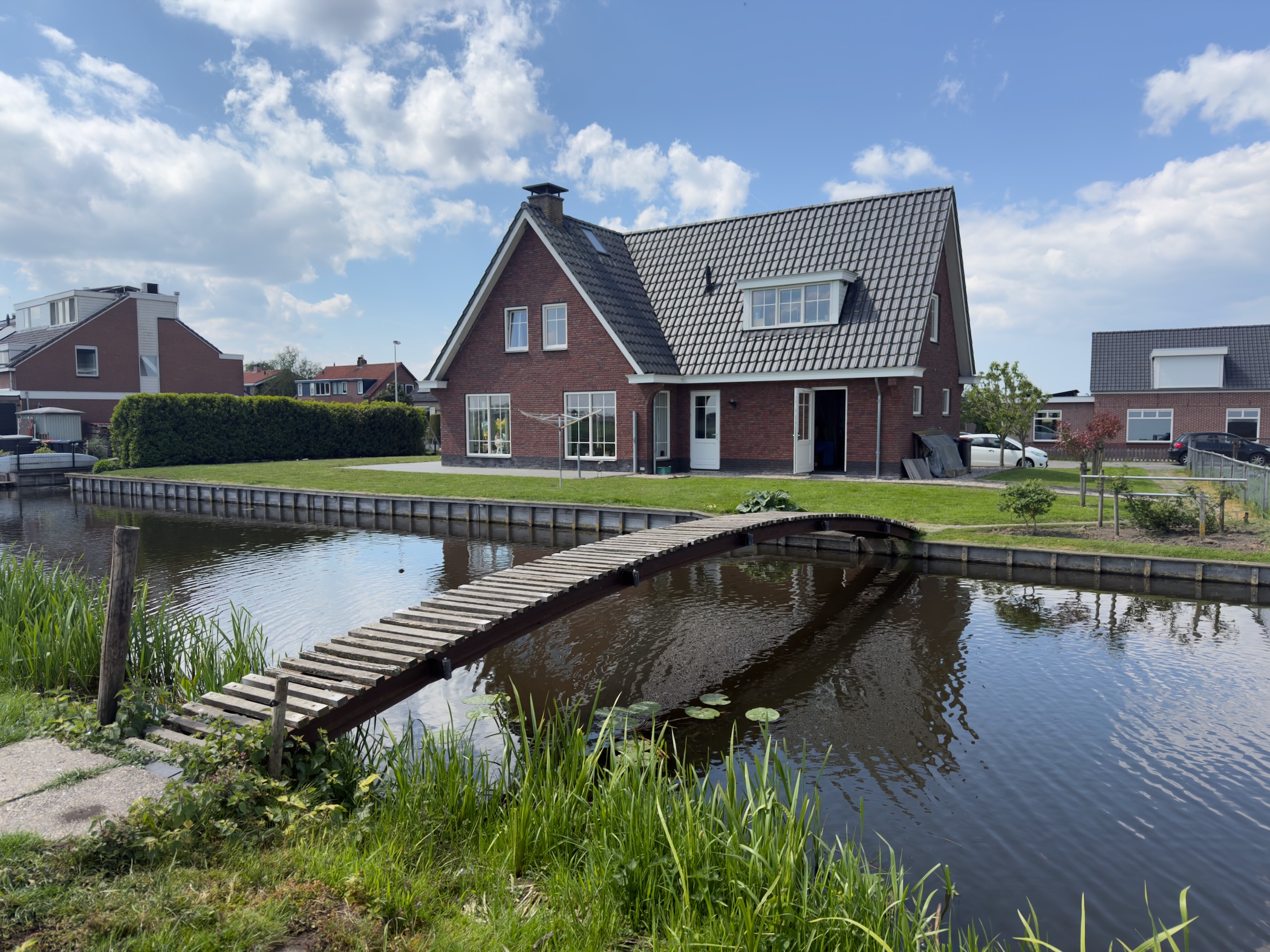Brick house with a steep dark roof beside a canal crossed by a narrow wooden footbridge