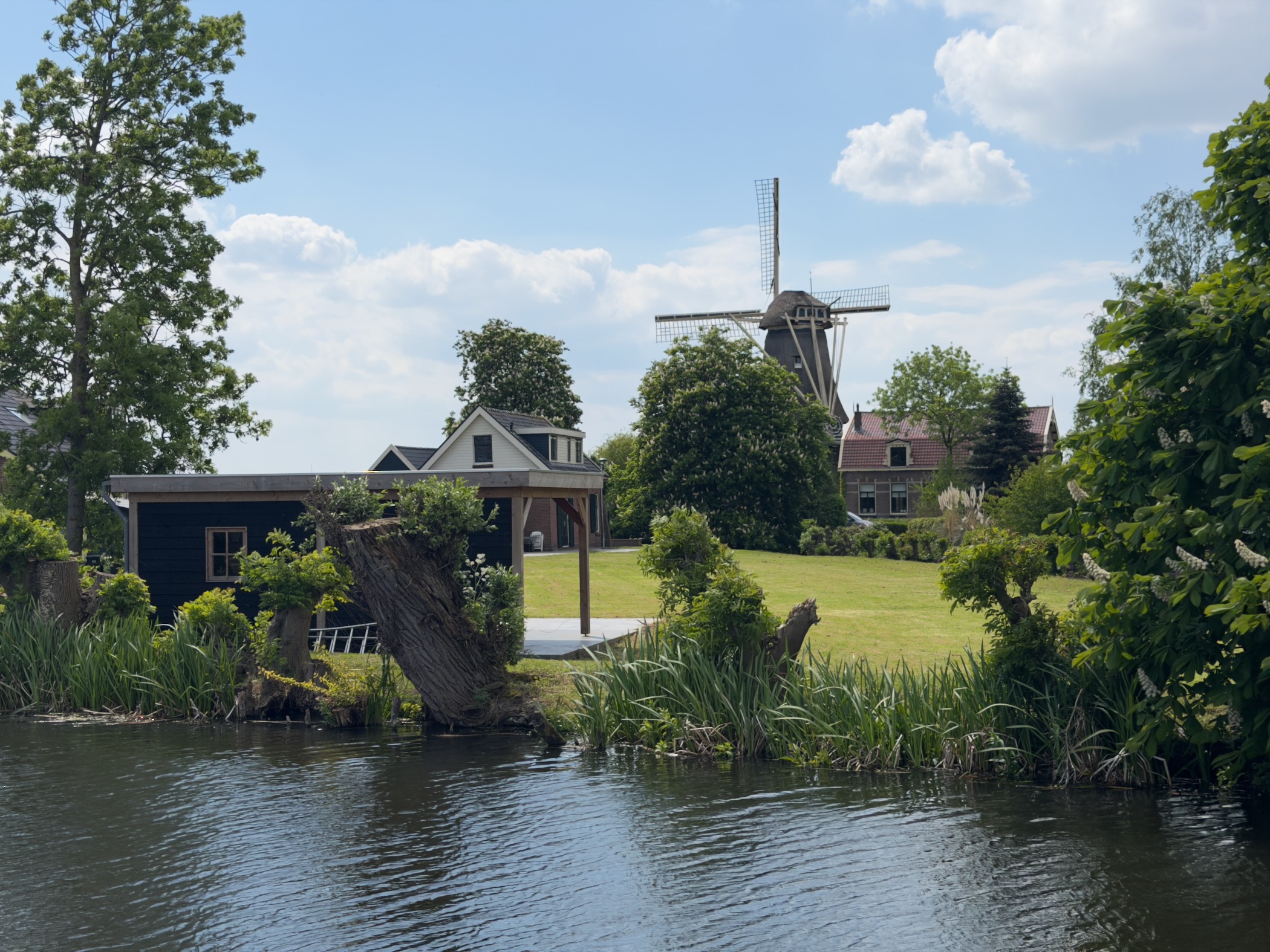 Tall white-capped windmill standing behind a small black-painted house and a calm canal