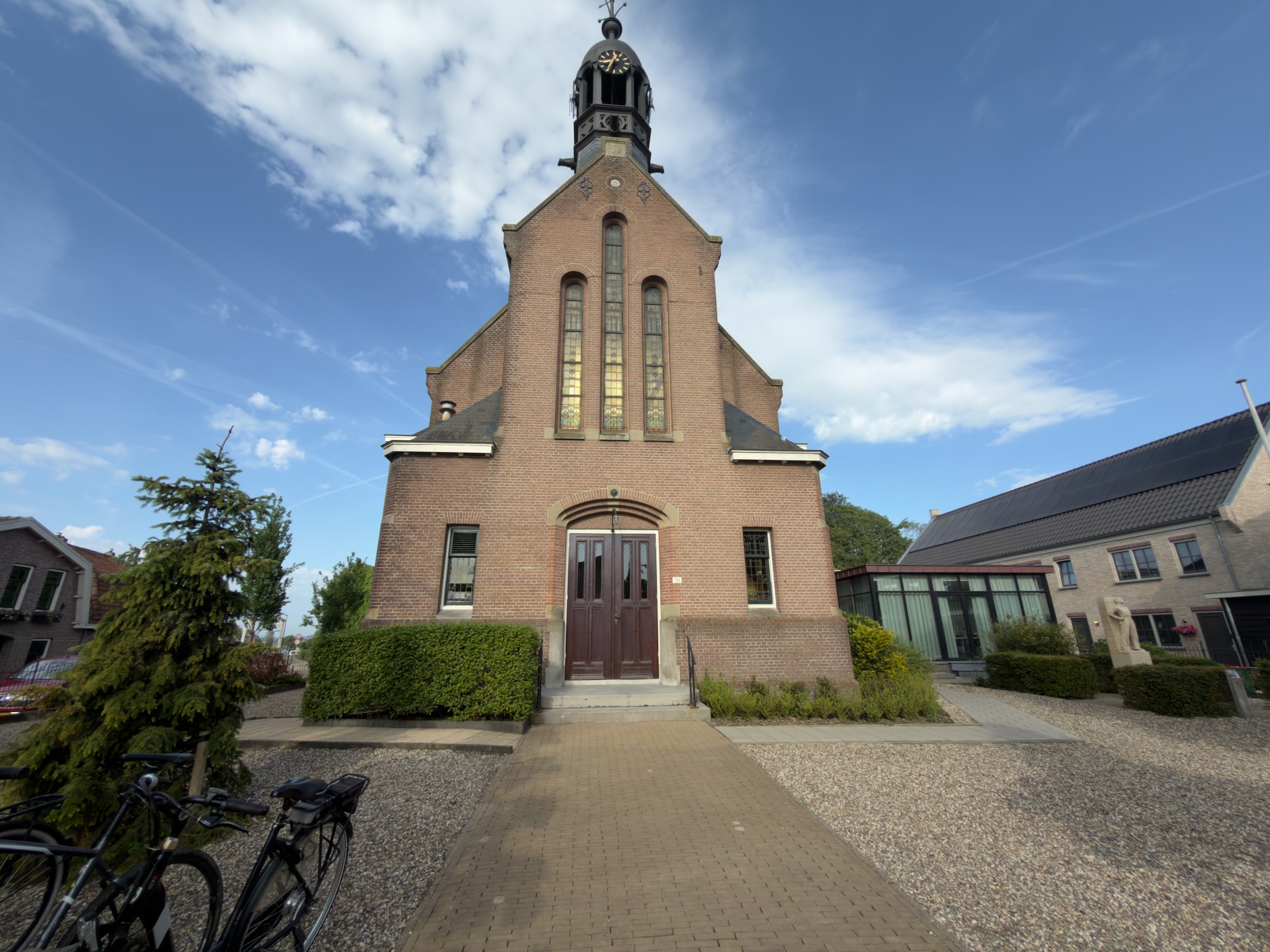 Brick village church with a tall stepped gable and small bell turret, bicycles parked at the front and a slender conifer to the left