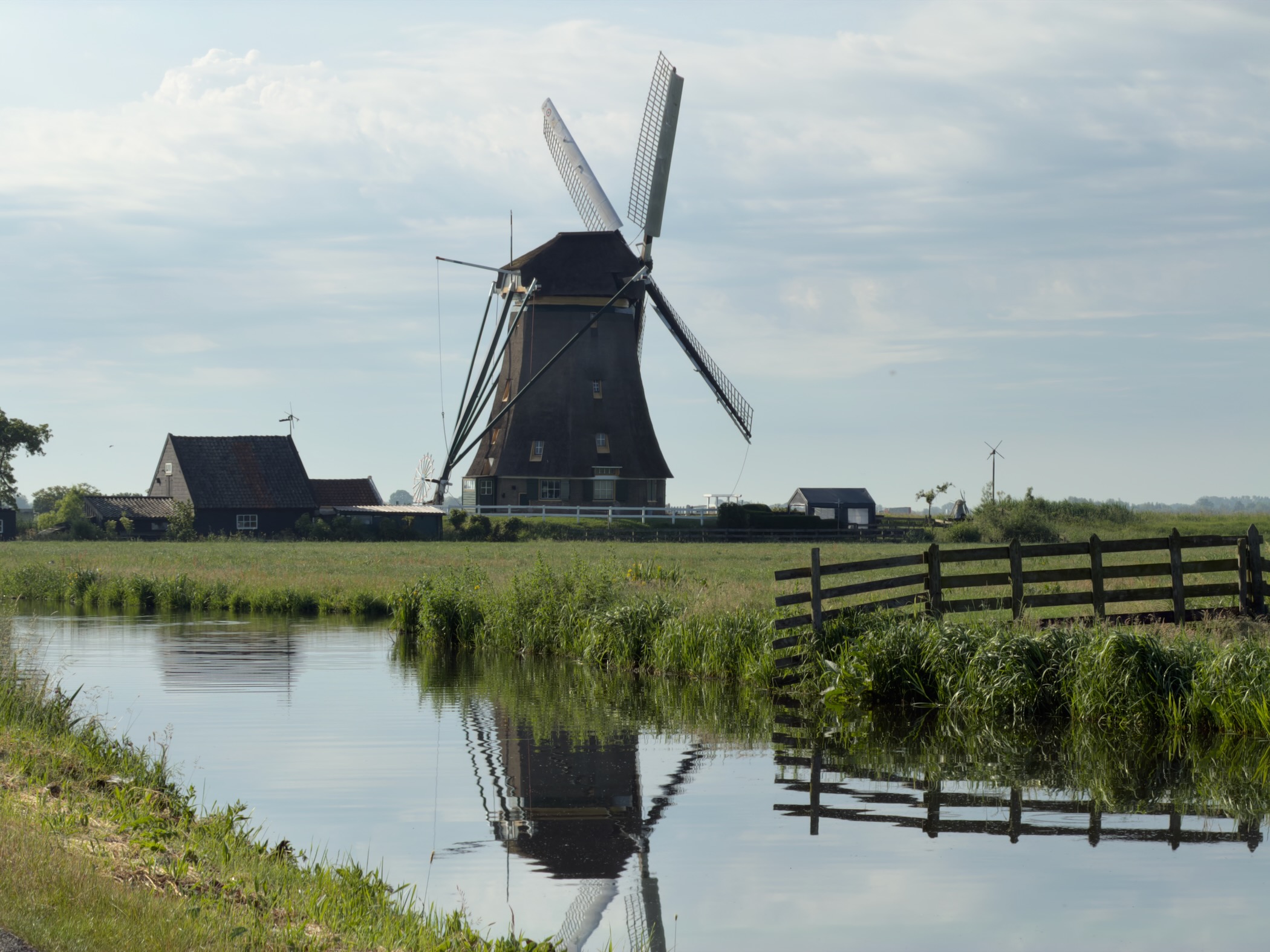 Tall dark thatched windmill with a small house beside it, reflected in a canal with a wooden fence in the foreground