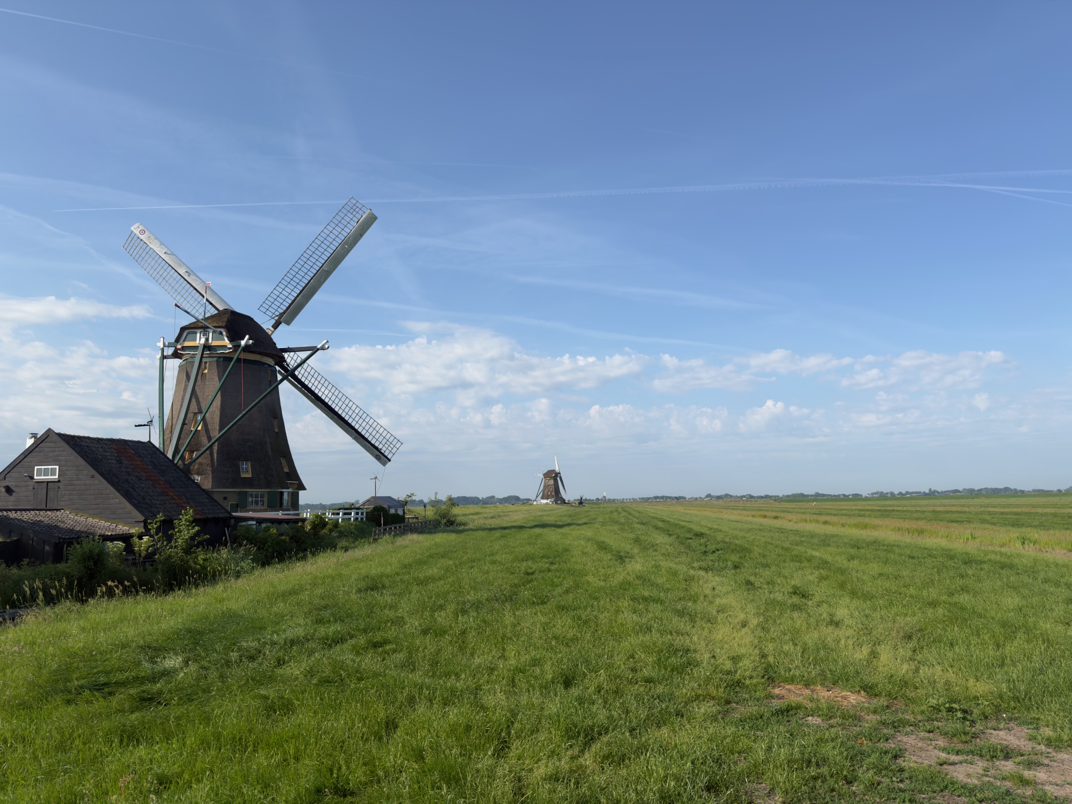 Thatched windmill with a house at its foot and a second windmill in the distance, beside a broad green dyke under a blue sky