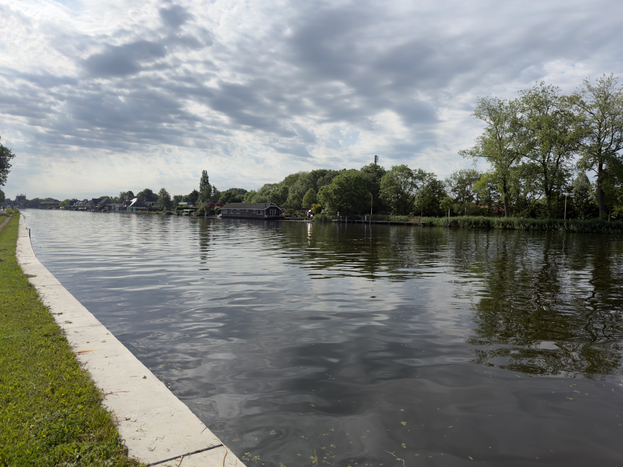 Broad tree-lined river with waterside houses under a cloudy sky, a paved path along the near bank