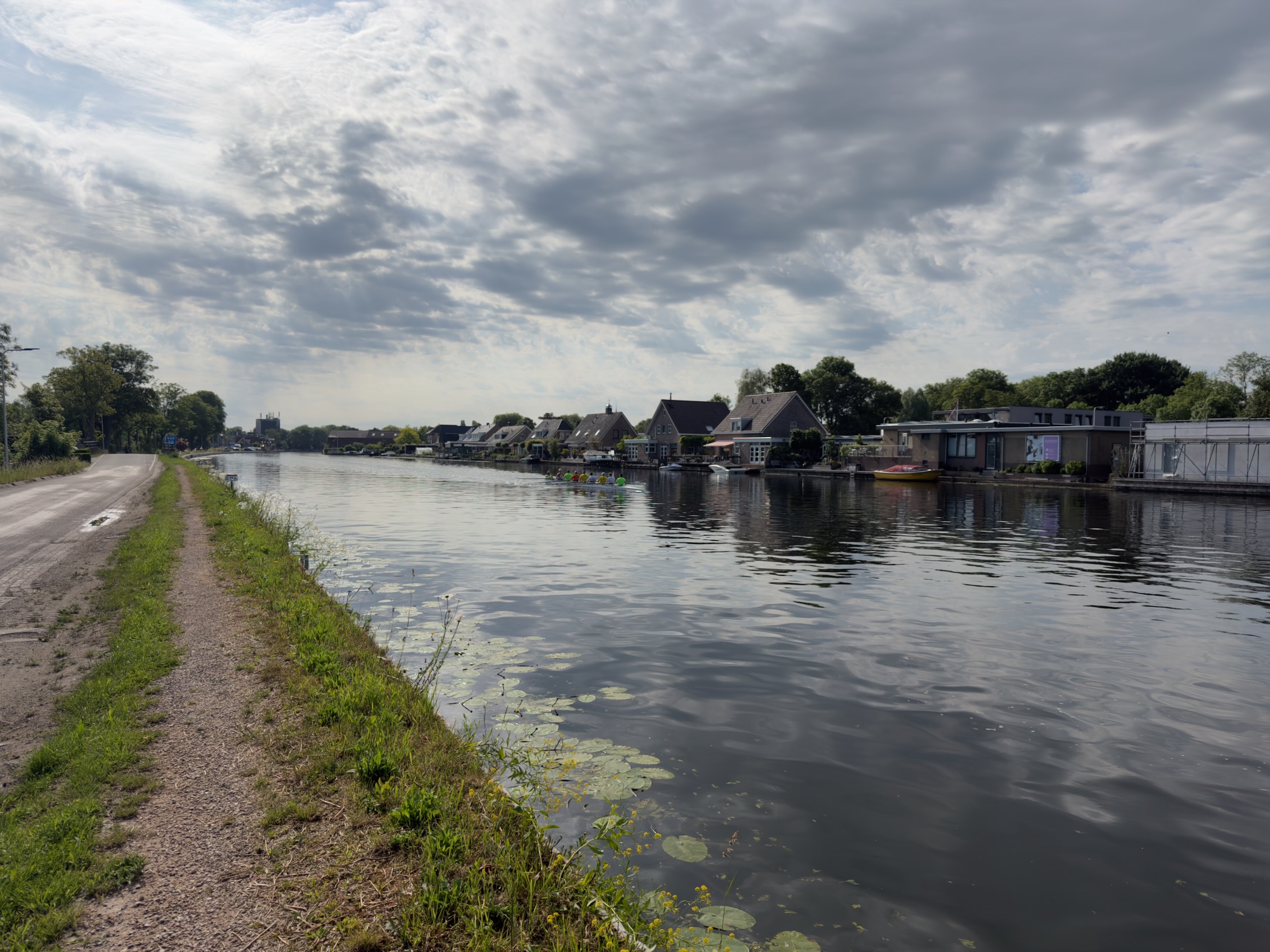 Wide canal flanked by houseboats and houses, a gravel track and grassy verge along the left bank under a cloudy sky