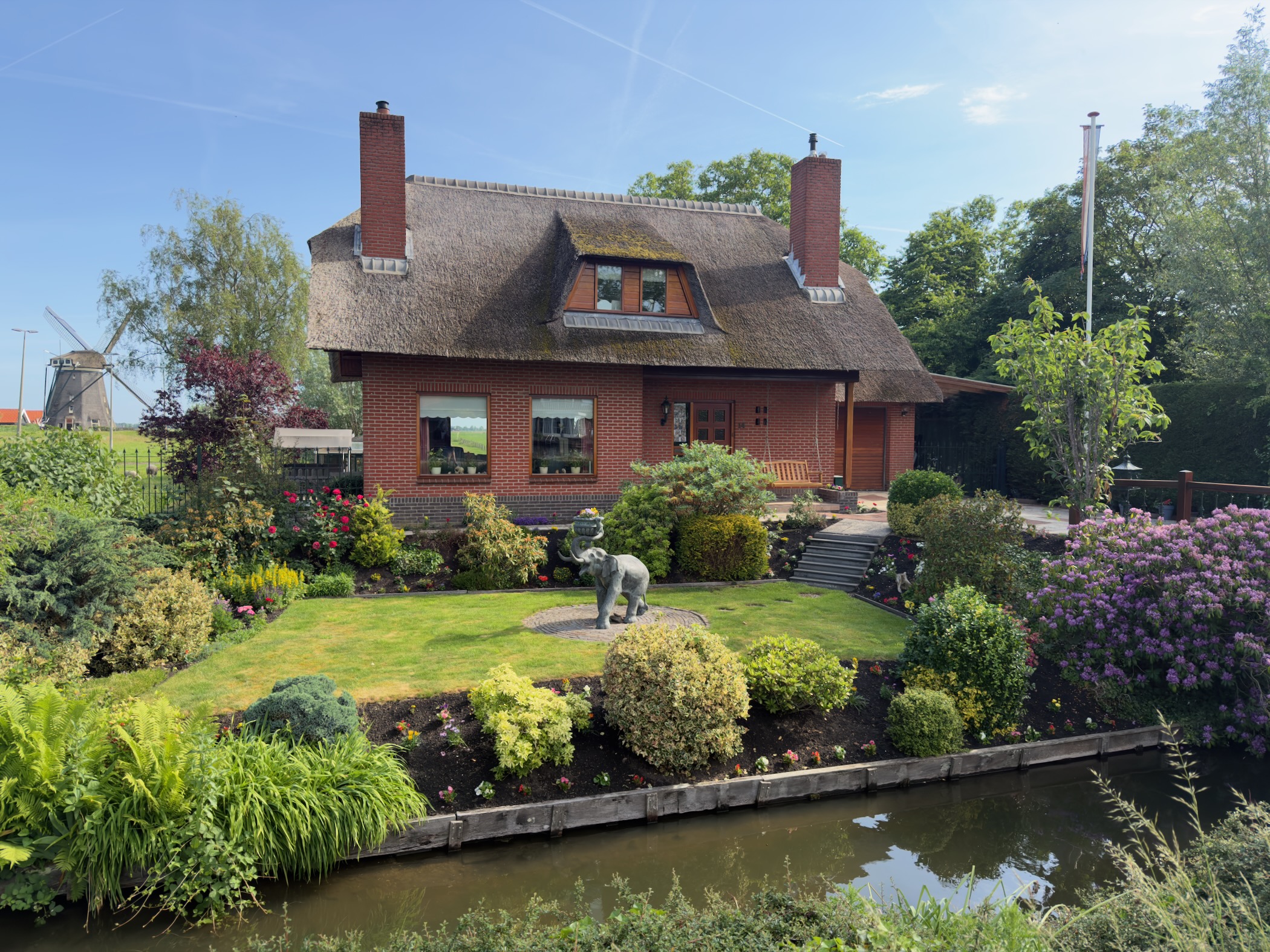 Brick house with a thatched roof and a bronze elephant statue in a colourful garden beside a canal, a windmill in the distance