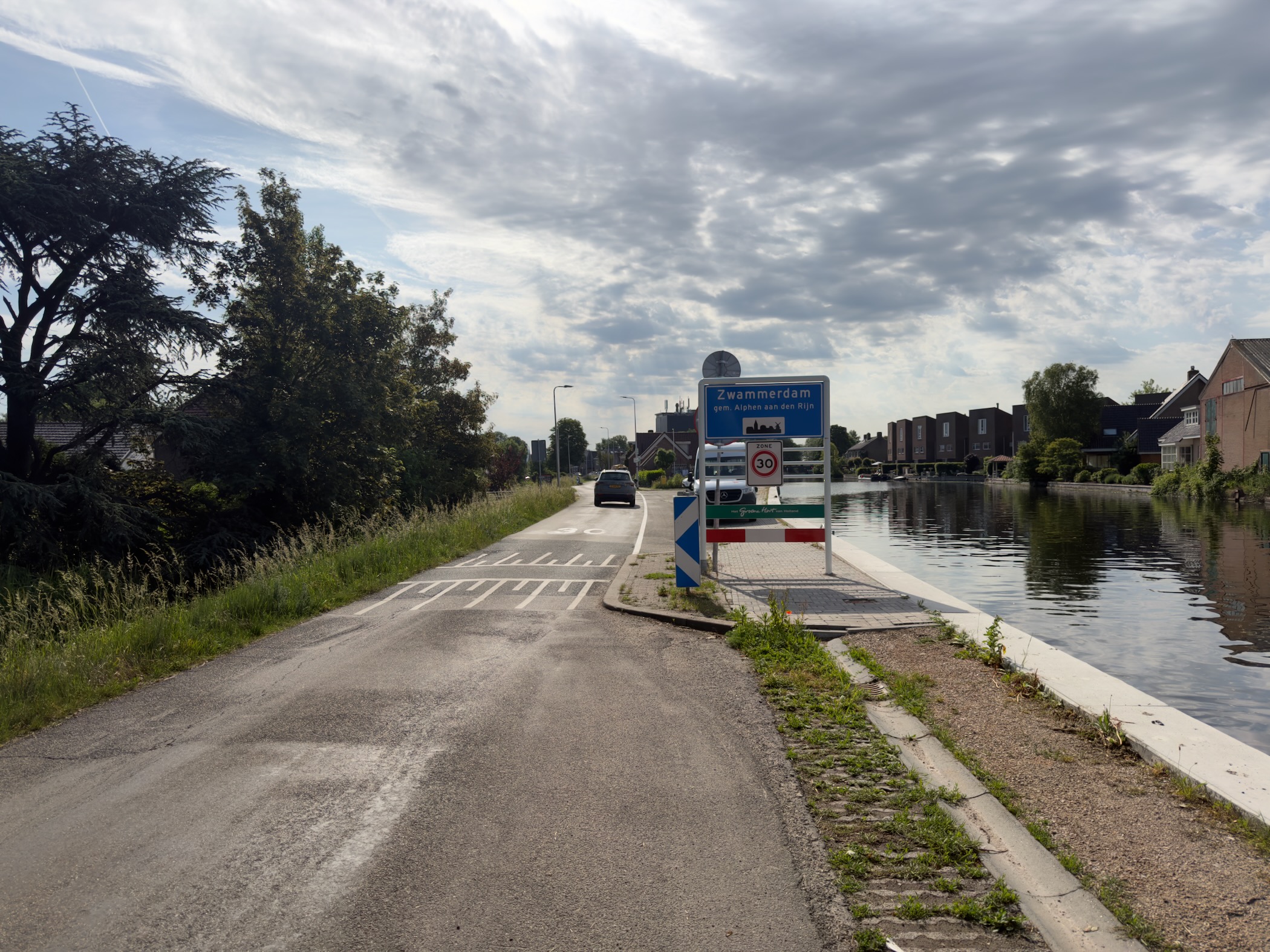 Road entering Zwammerdam past the town name sign, with a canal and houses to the right under a grey sky