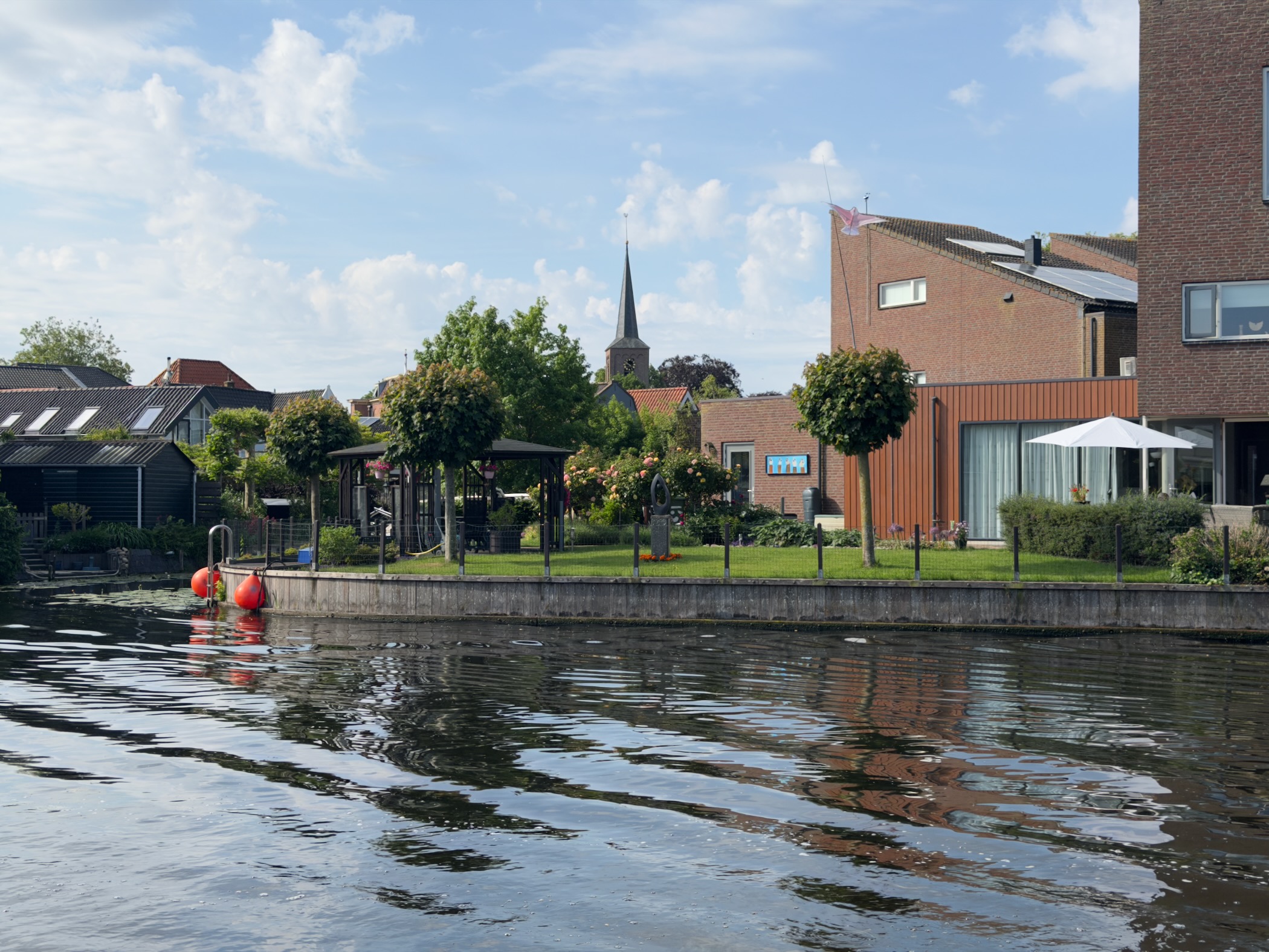 Waterside gardens and houses across a canal with a church spire rising behind, red mooring buoys near the bank
