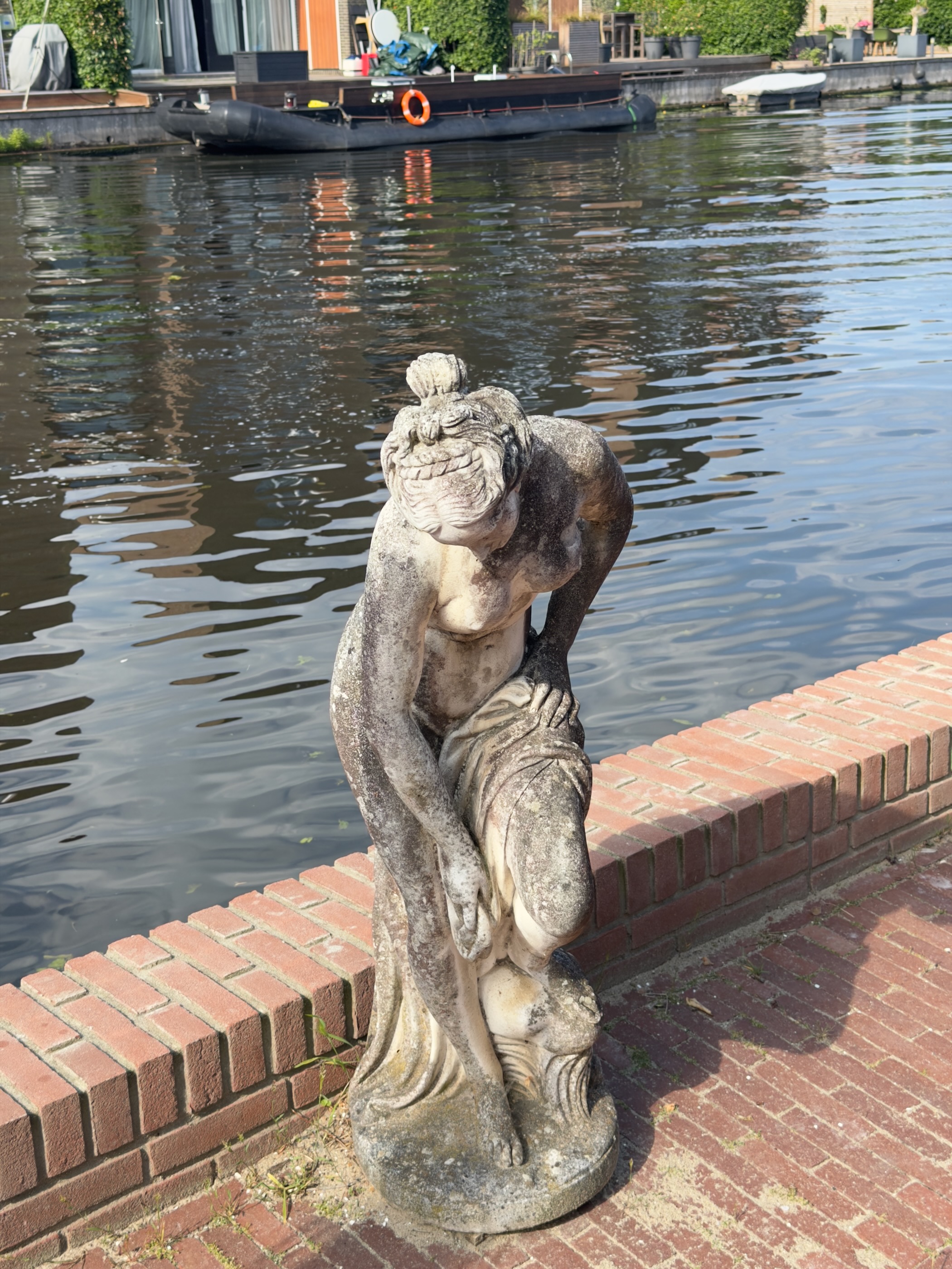 Weathered stone statue of a crouching woman on a brick quay beside a canal, a moored barge behind