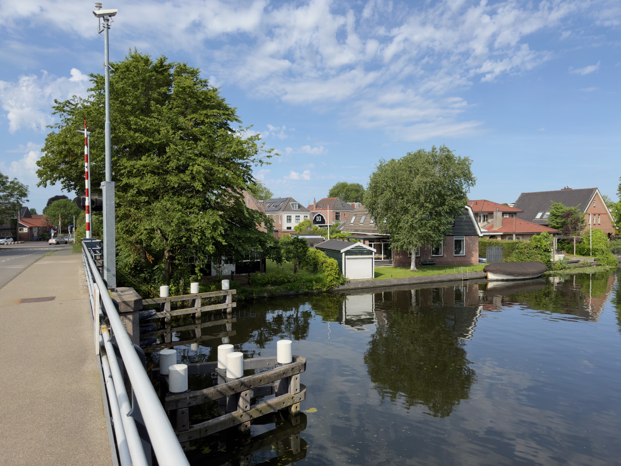 Canal lined with houses and trees seen from a bridge railing, mooring posts and a small boat along the far bank