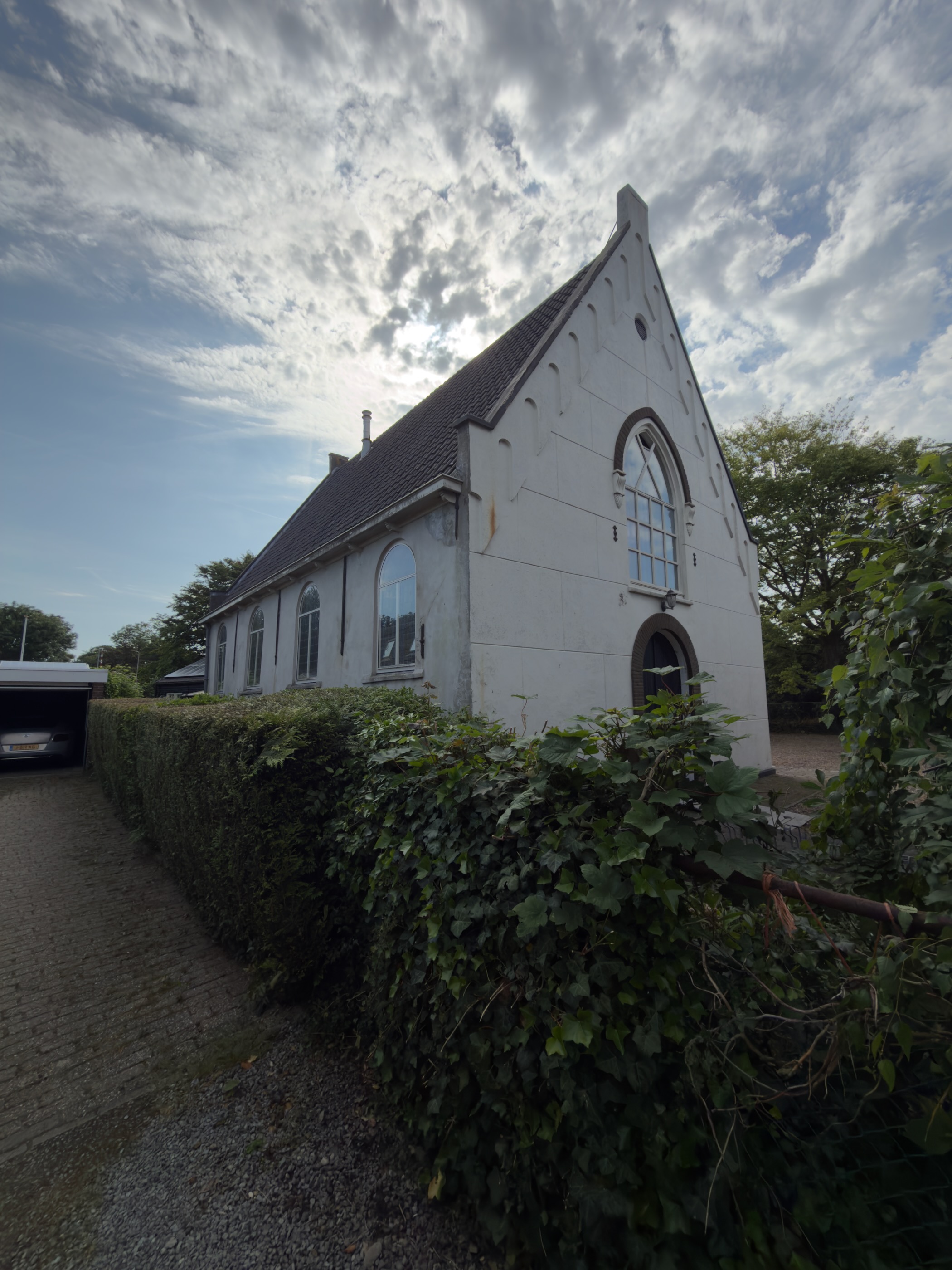 White-painted former church with a stepped gable and arched windows behind an ivy-covered hedge, a car parked to the left
