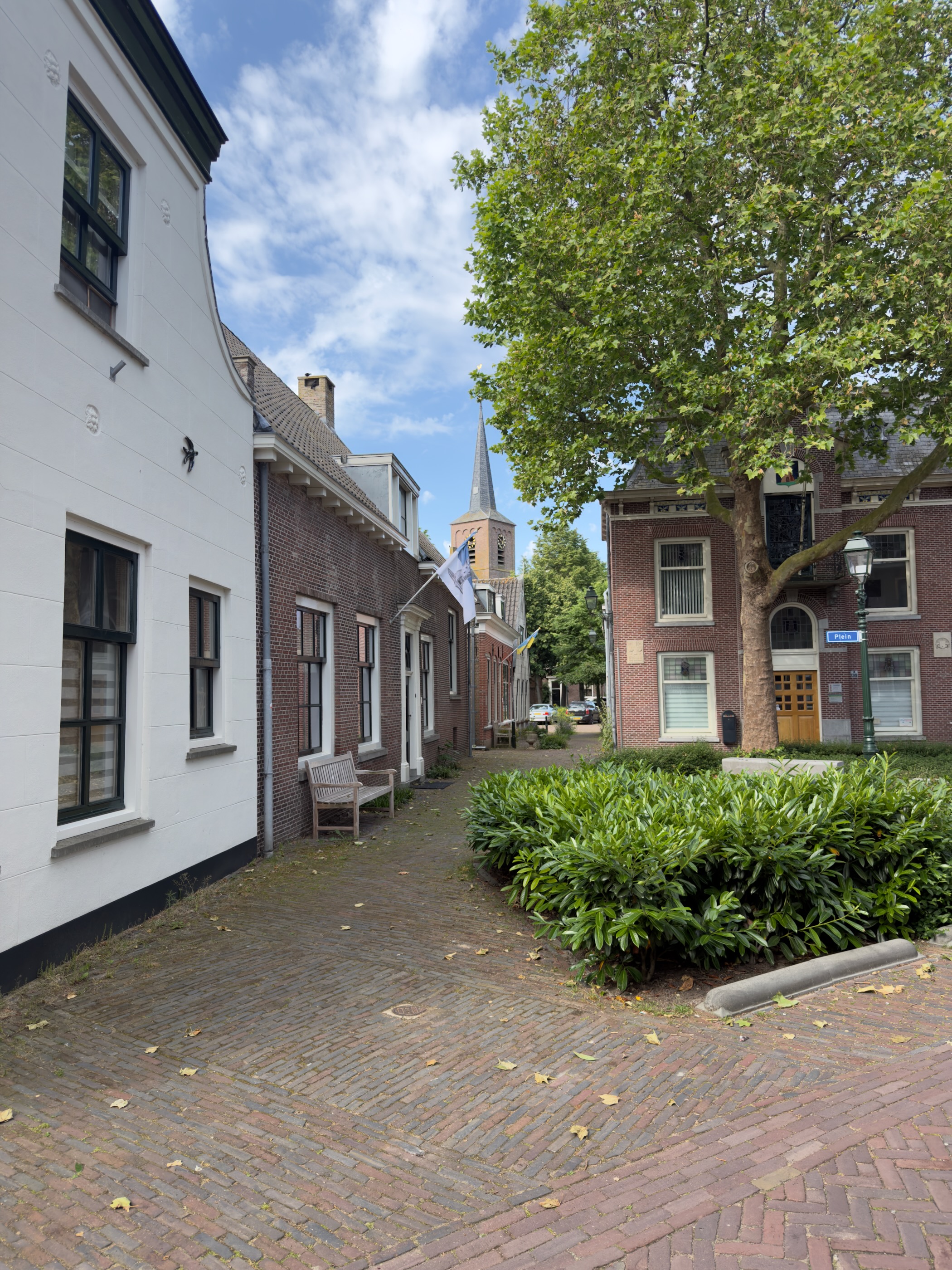 Quiet brick-paved village street with white and brick houses, a bench, and a church spire rising at the end