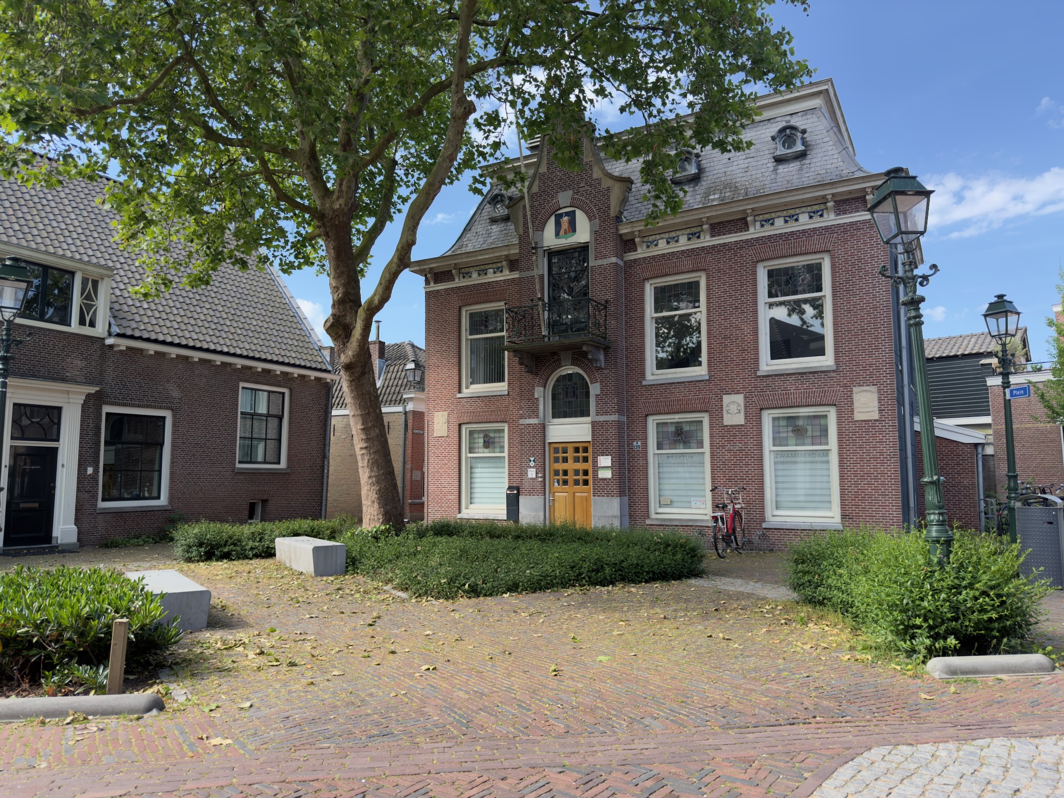 Grand brick town house with a stepped gable and ornate dormer, set behind a tree on a brick-paved square