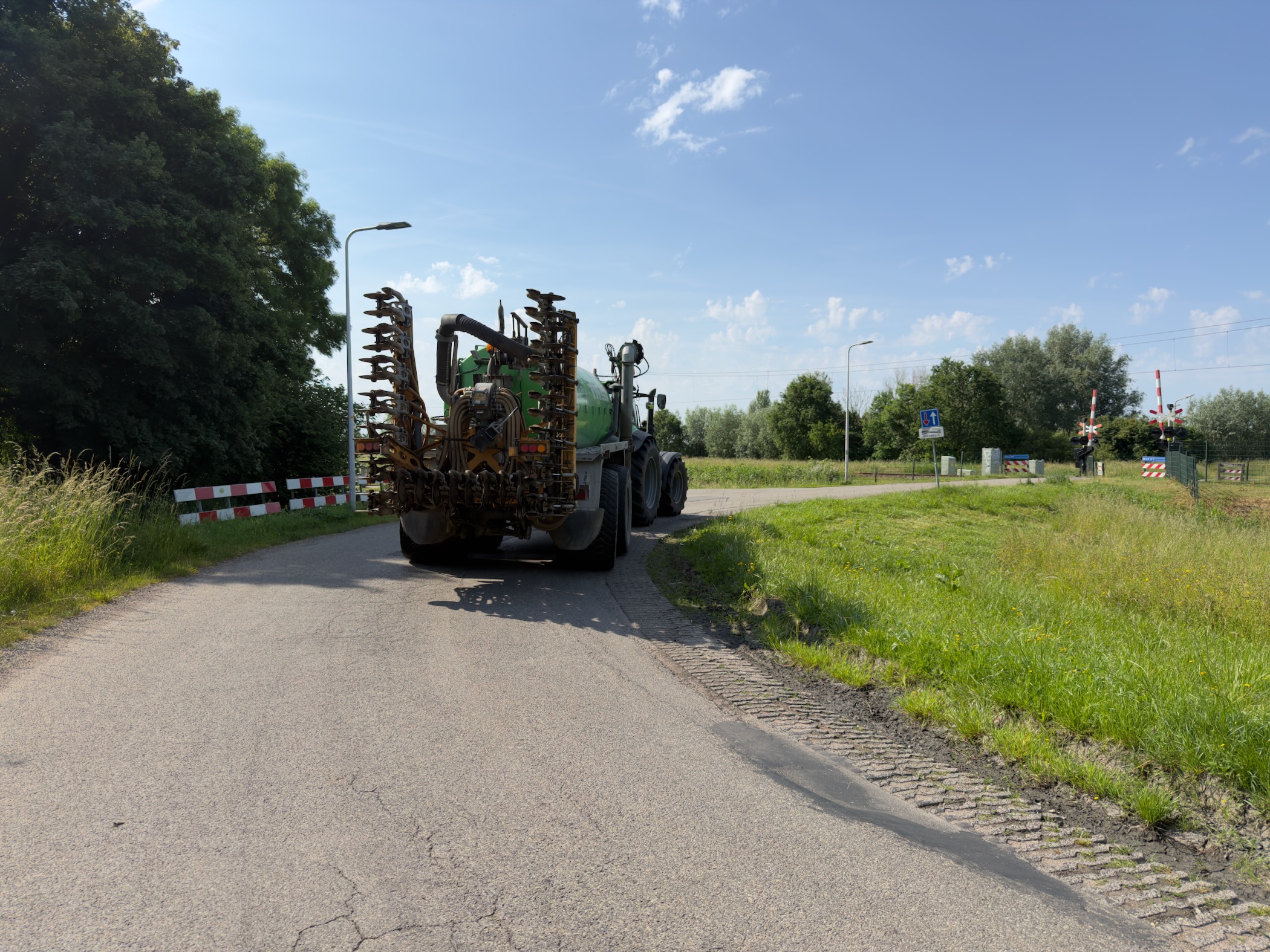 Green slurry tanker with a folded implement parked on a country lane beside a railway crossing