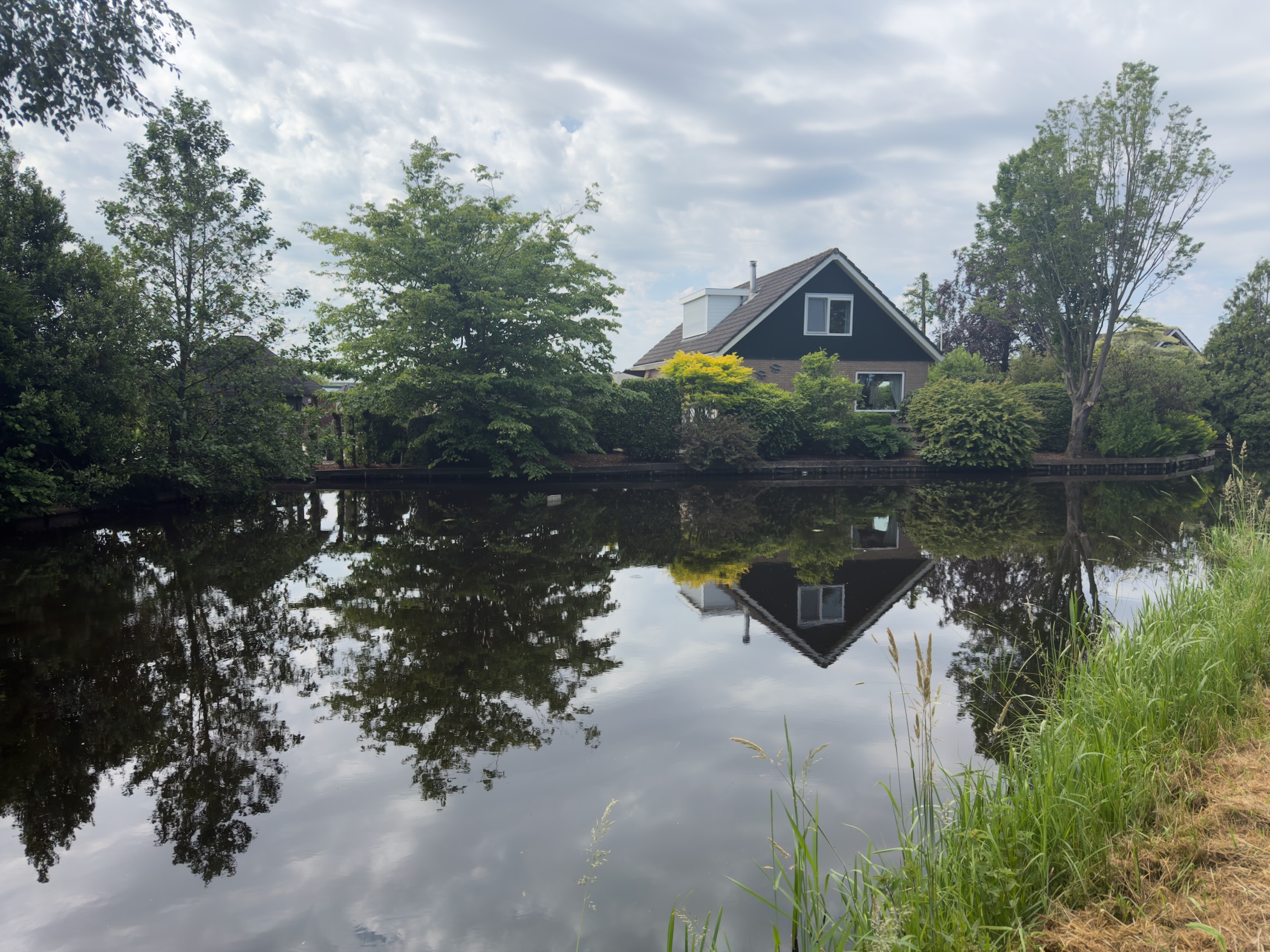 Dark-clad house with a steep roof among trees on the far bank of a calm canal, mirrored in the still water under a cloudy sky