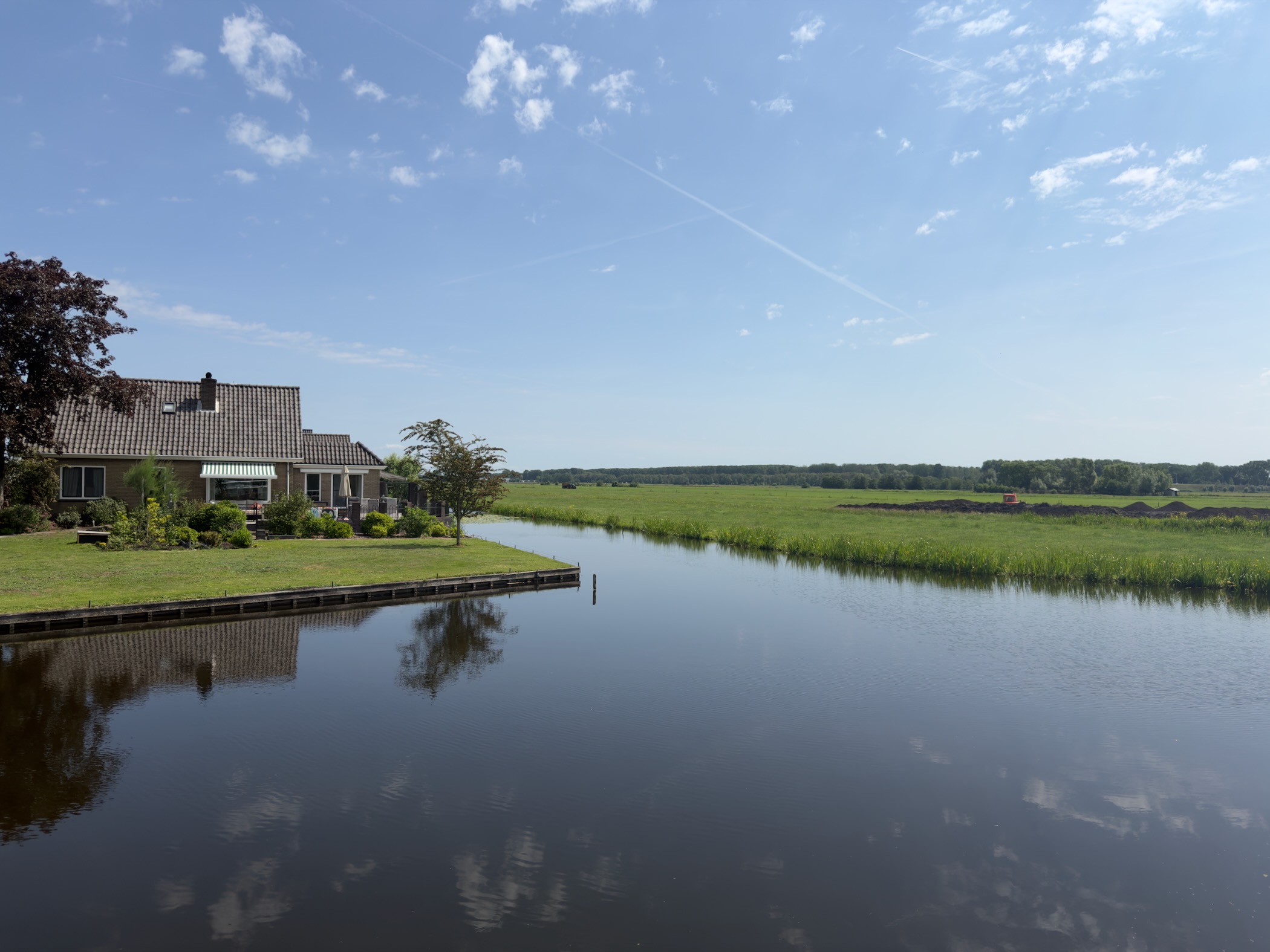 House with a tiled roof and garden on the left bank of a broad canal, open polder and a distant tree line beyond under a blue sky