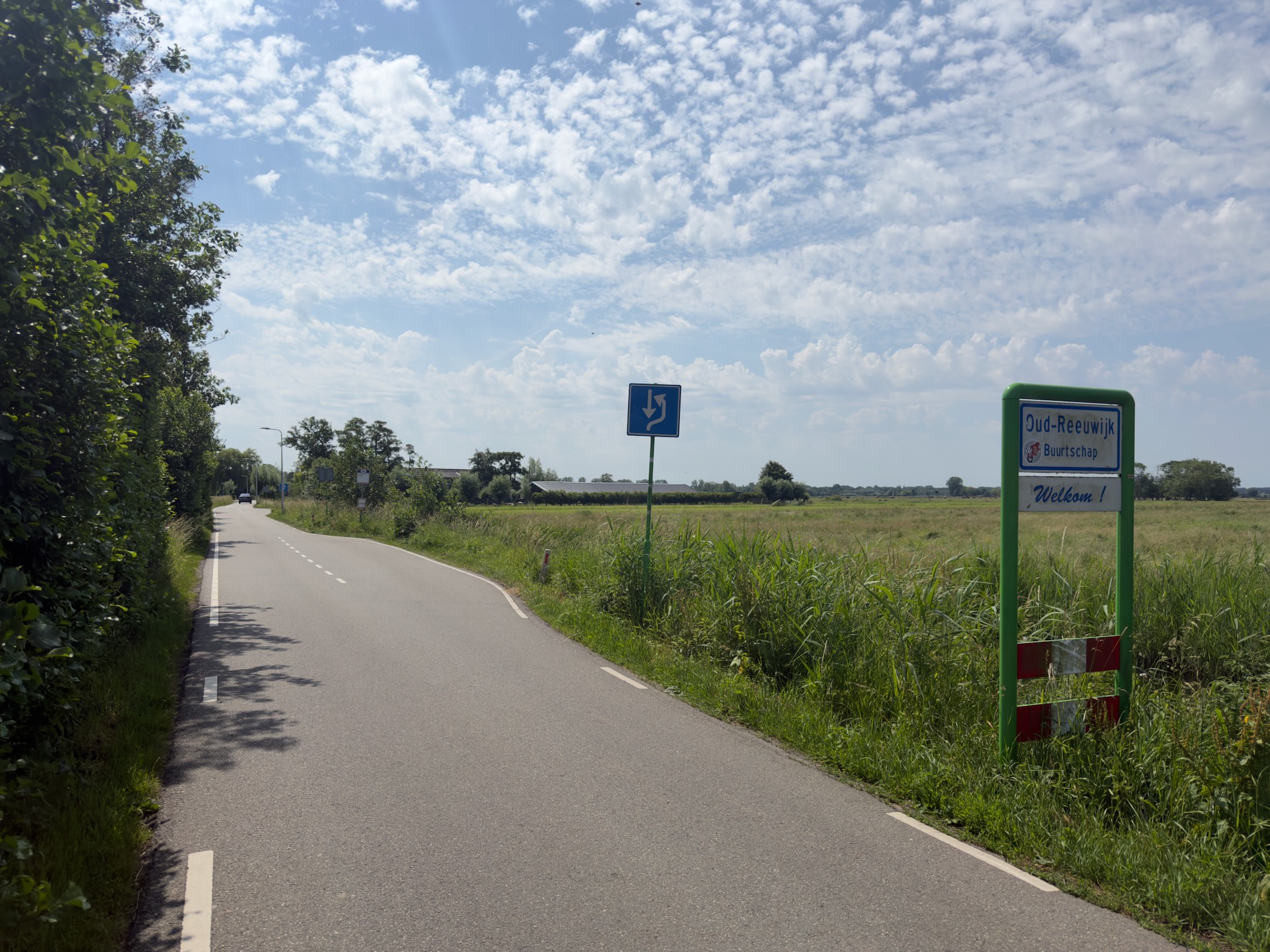 Country road entering Oud-Reeuwijk past the green town name sign, with open meadow on the right under a partly cloudy sky