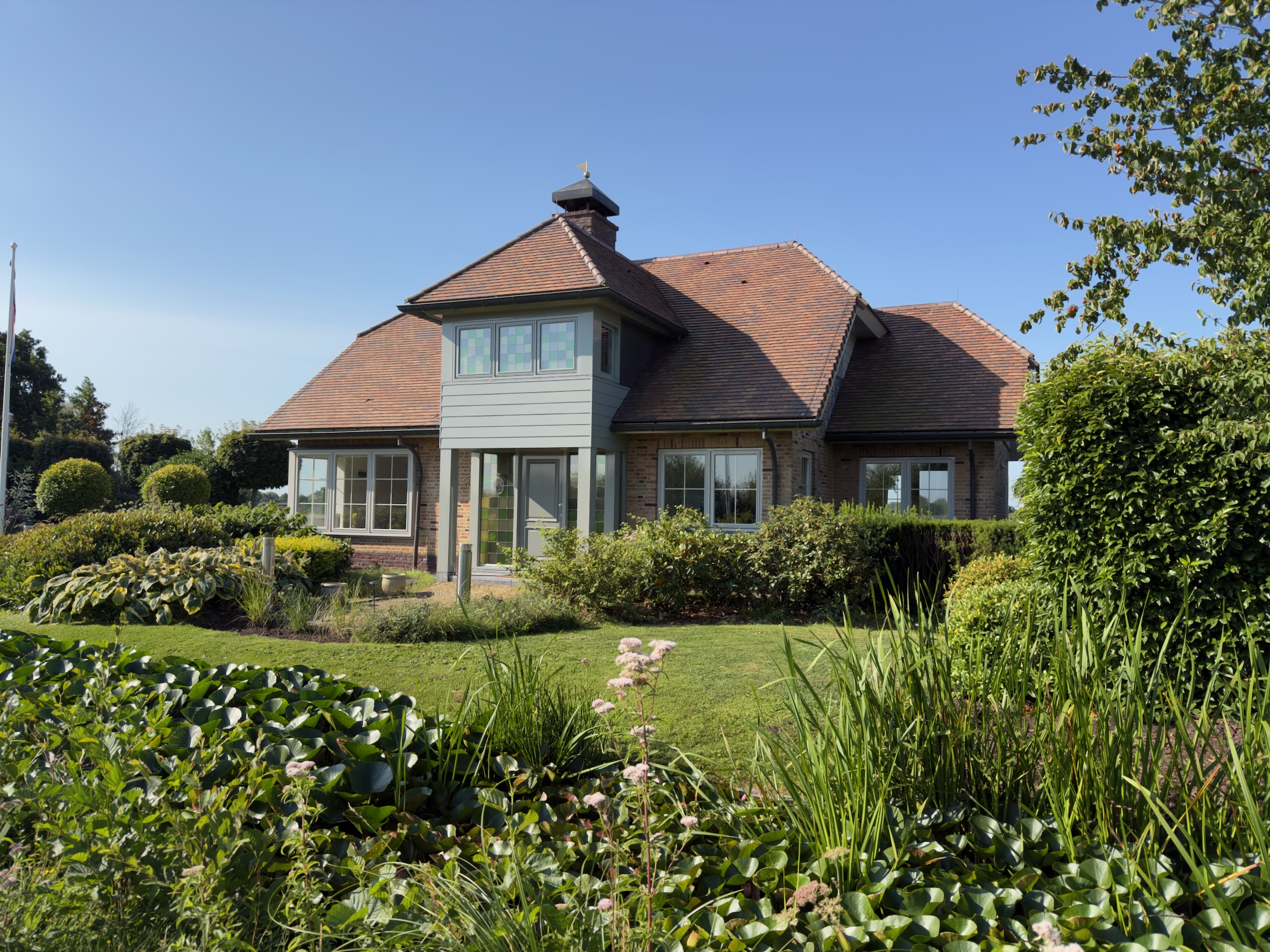 Large brick house with a red tiled roof and grey timber bay, set in a manicured garden with hostas and a lily pond