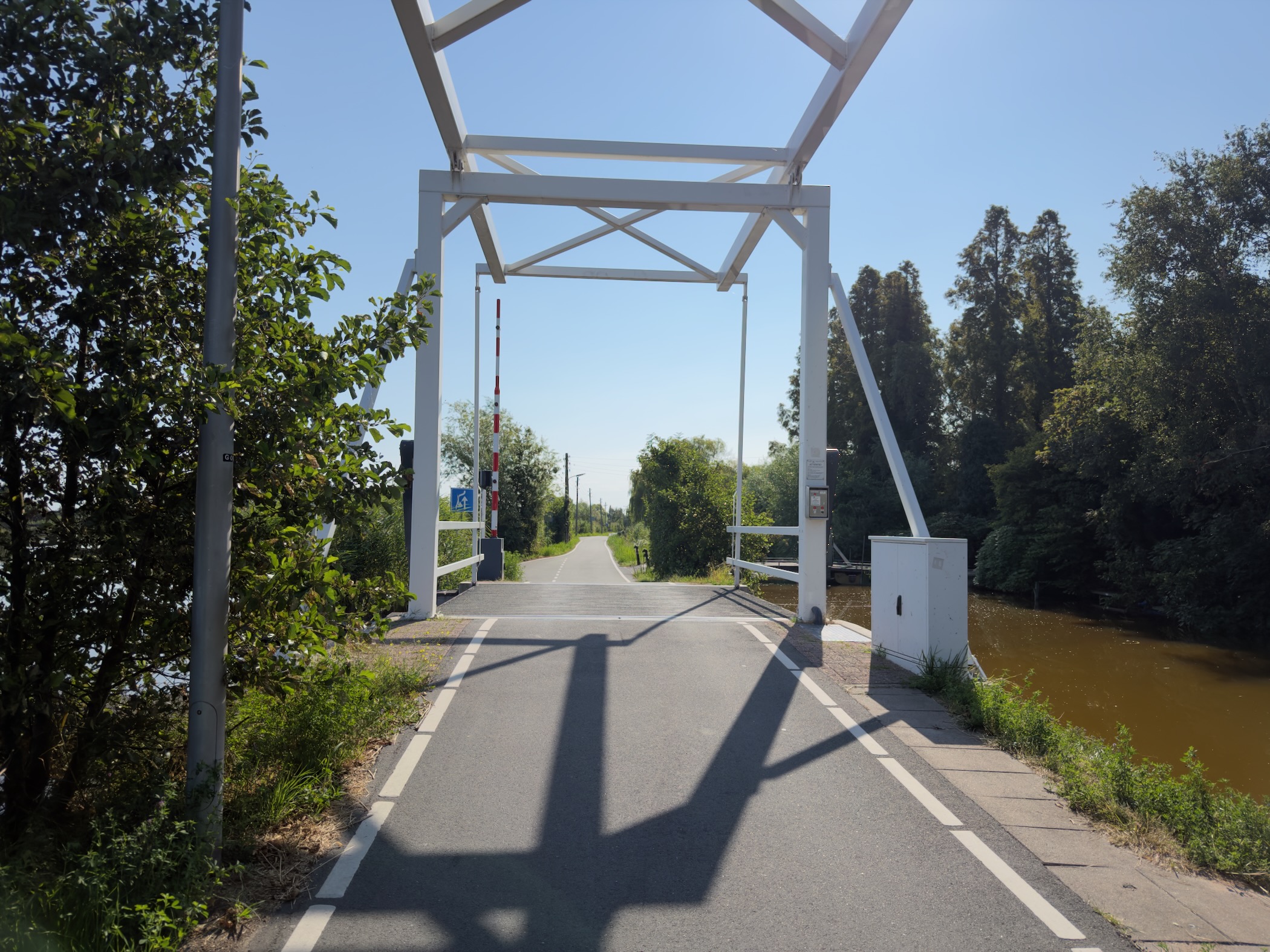 White steel bascule bridge over a canal with long shadows of the truss cast across the road deck