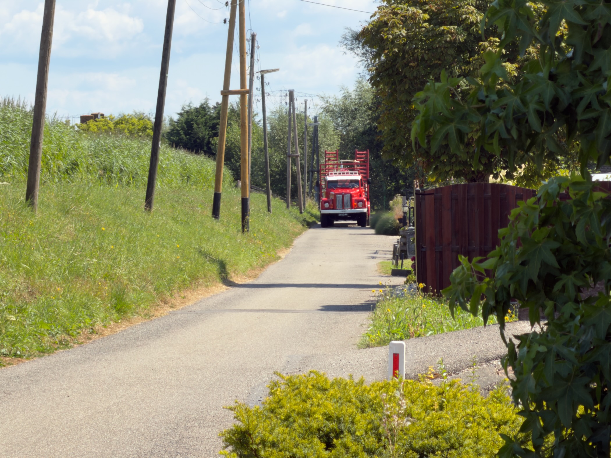 Narrow country lane with a bright red truck approaching between a grassy bank and garden fences