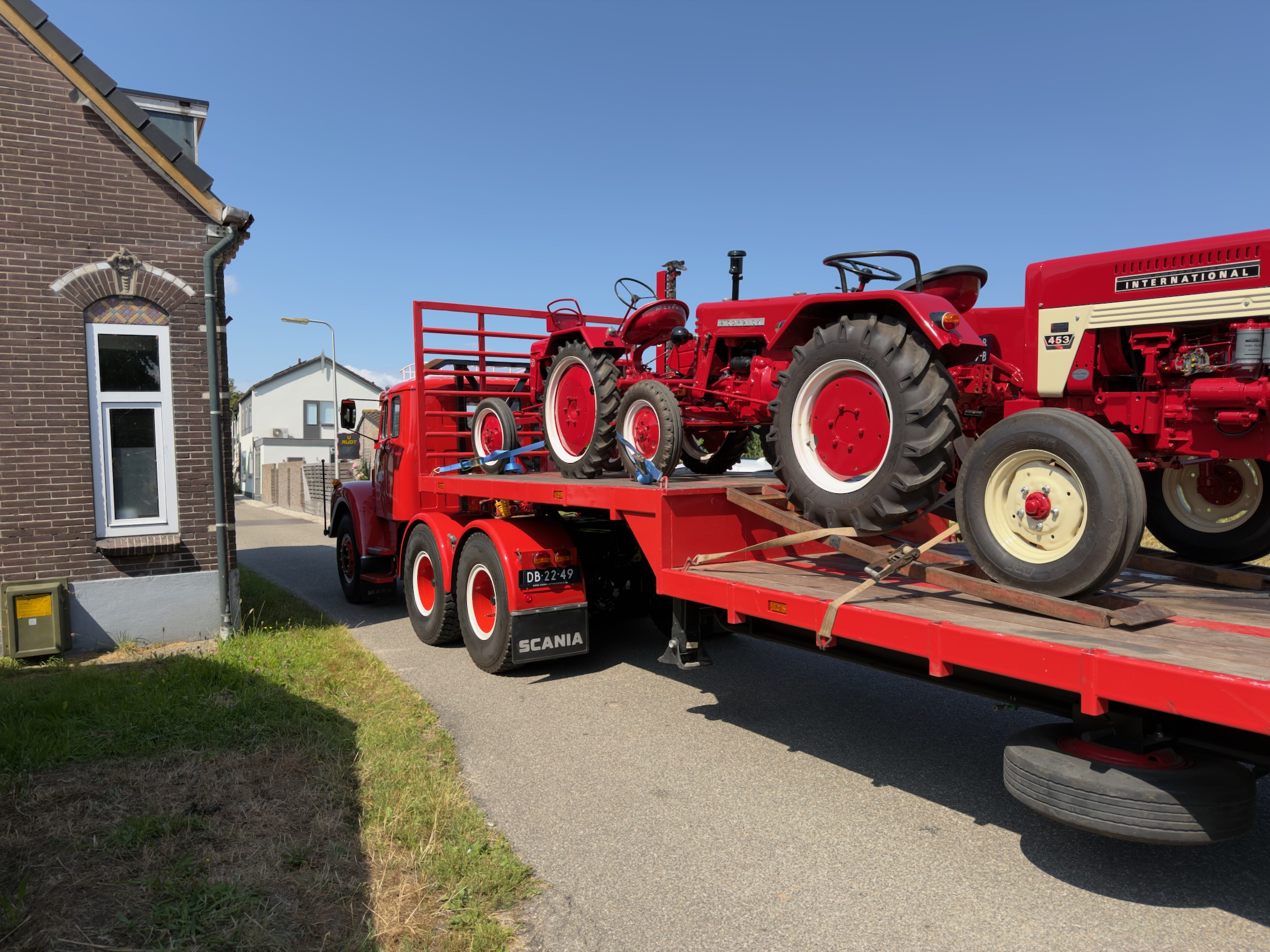 Vintage red Scania flatbed truck carrying classic red International tractors along a village street