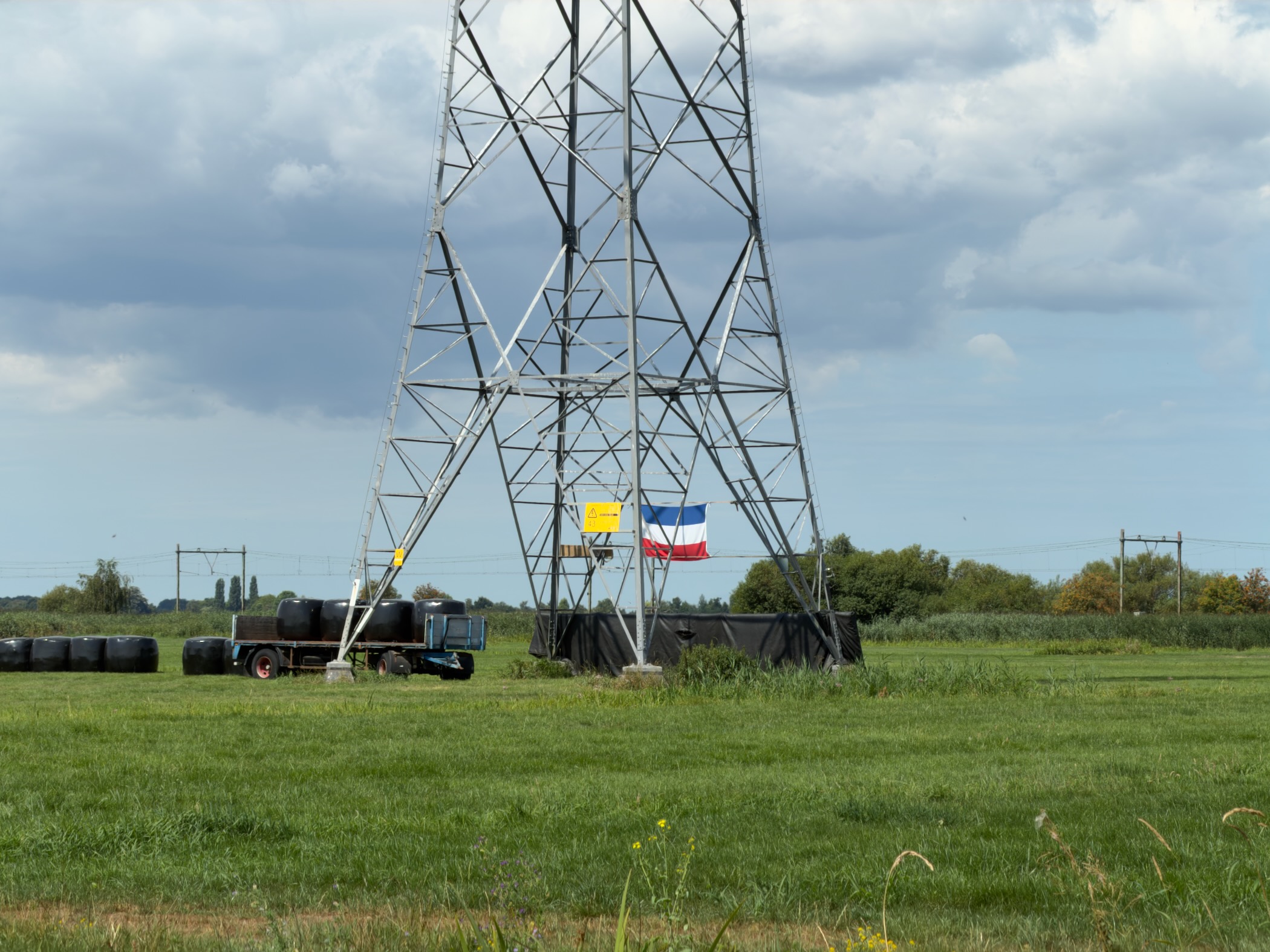Steel electricity pylon in a green field with black silage bales, a trailer, and a French flag hanging from the frame