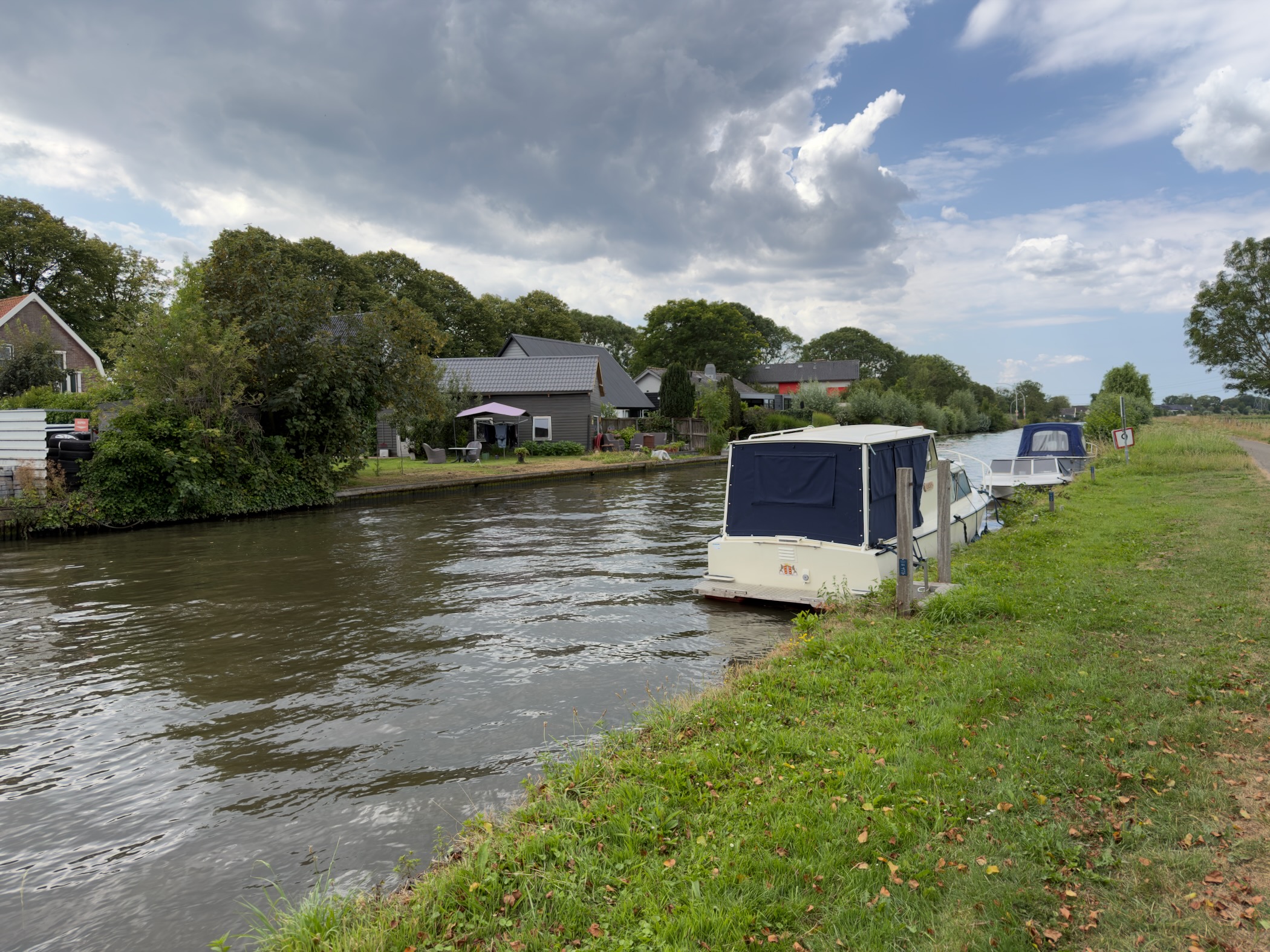 Canal with white cabin boats moored along a grassy bank and houses under a sky of dark and white cloud