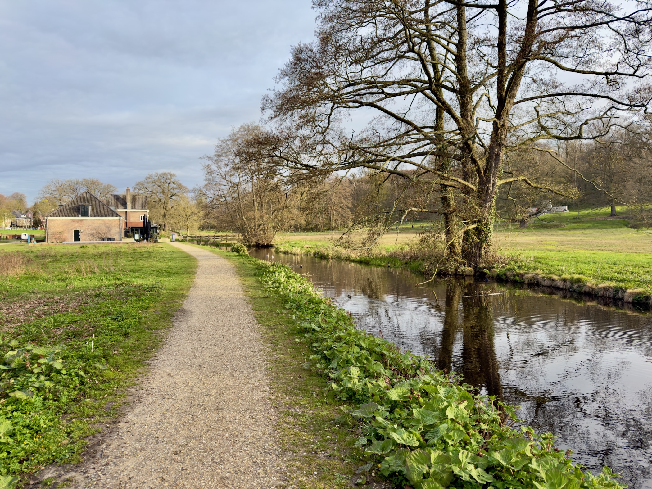 Gravel path along a stream with trees and a farm building in Sonsbeek park