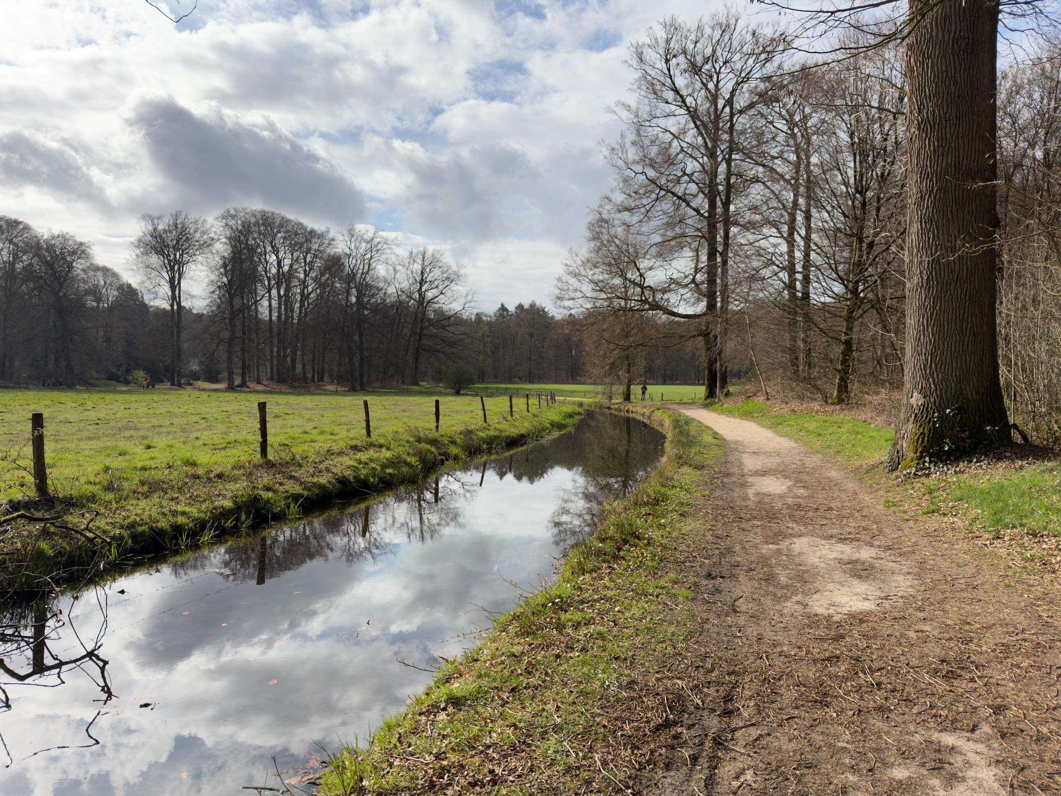 Path along a calm stream reflecting clouds with fenced meadows